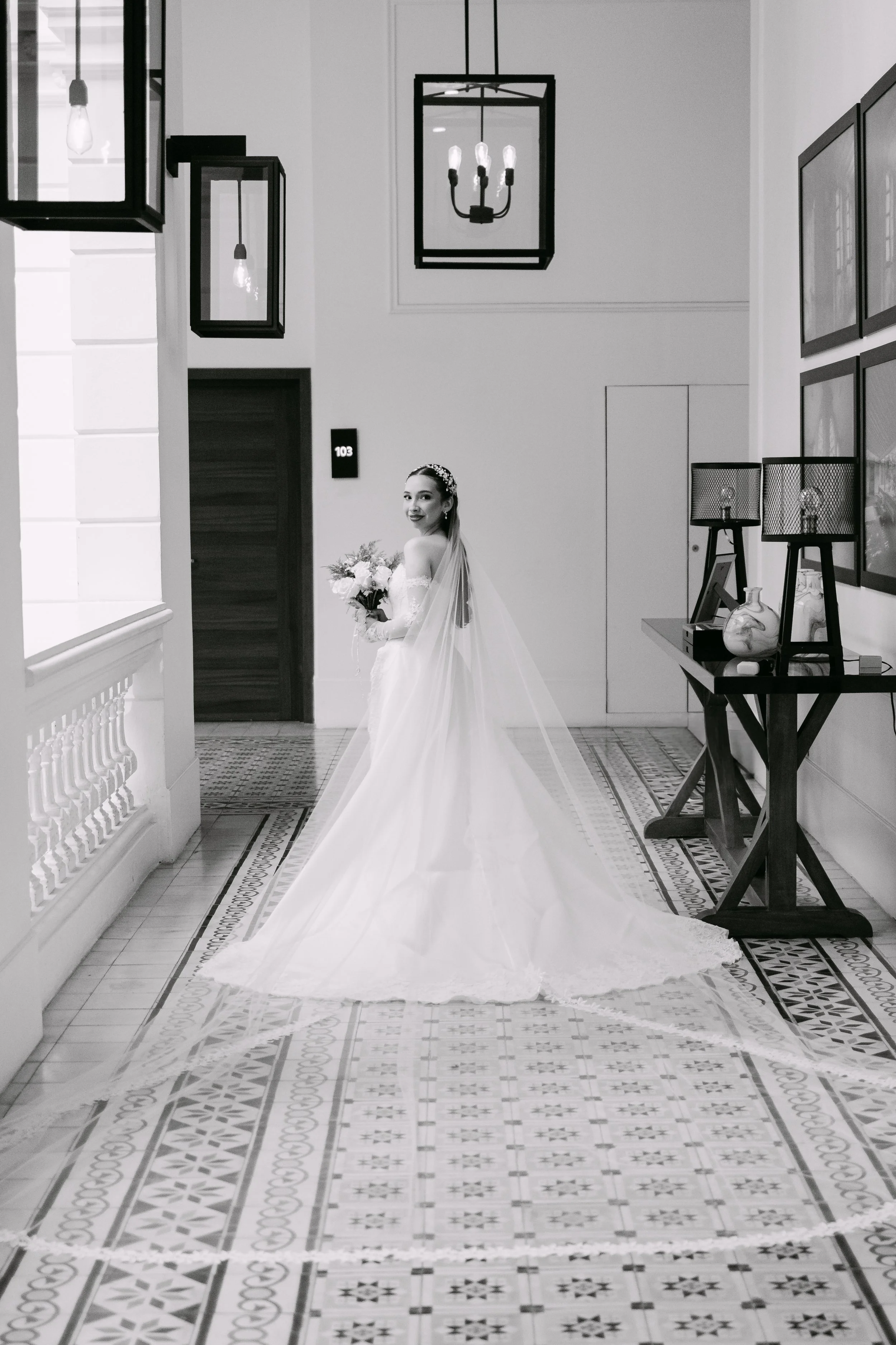 Mujer vestida de novia sosteniendo un ramo de flores en un pasillo de estilo moderno y elegante, con decoración minimalista y cuadros en la pared.