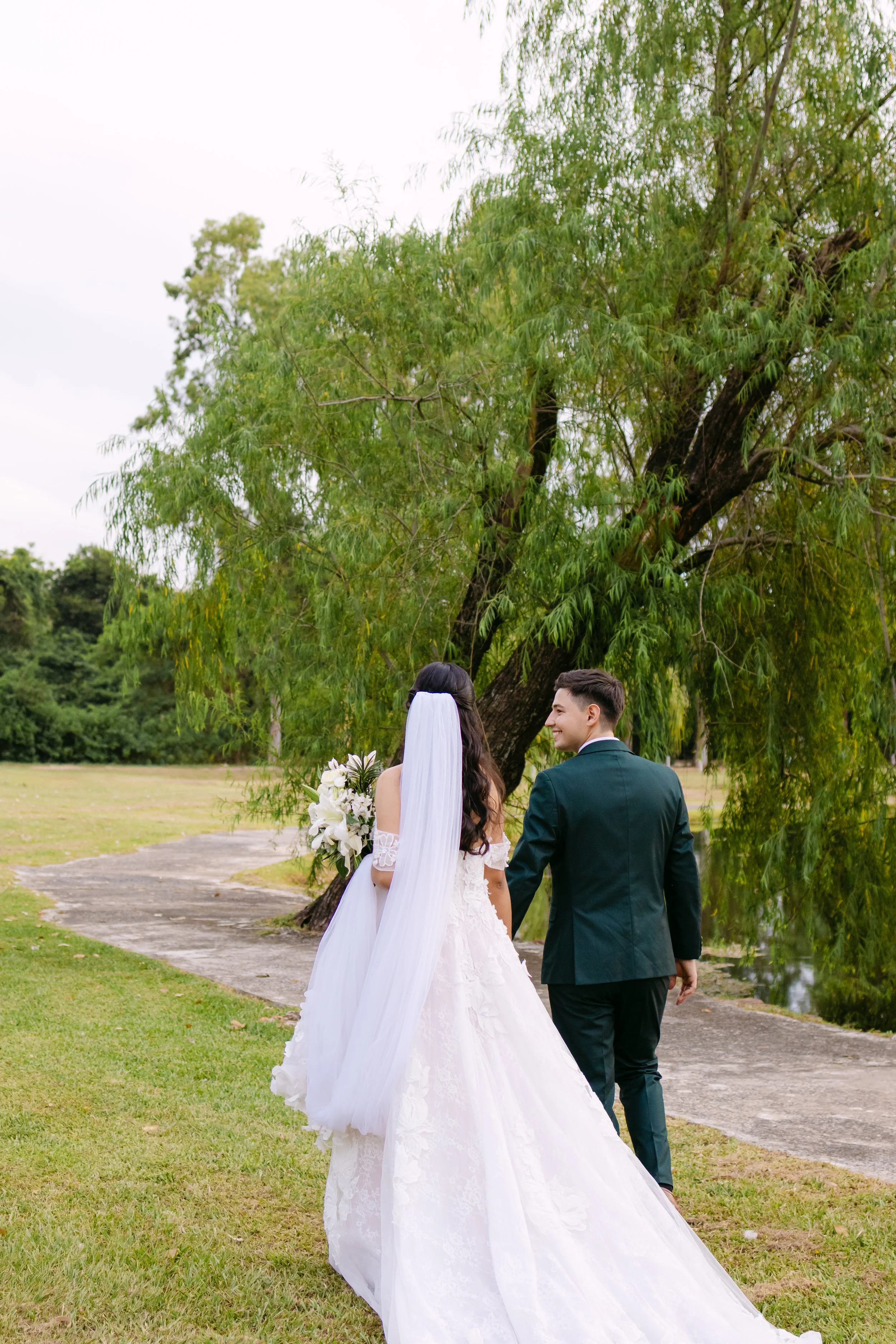 Una pareja de boda caminando por un parque verde, la mujer lleva un vestido de novia y el hombre un traje formal. Ella sostiene un ramo de flores blancas y ambos sonríen.