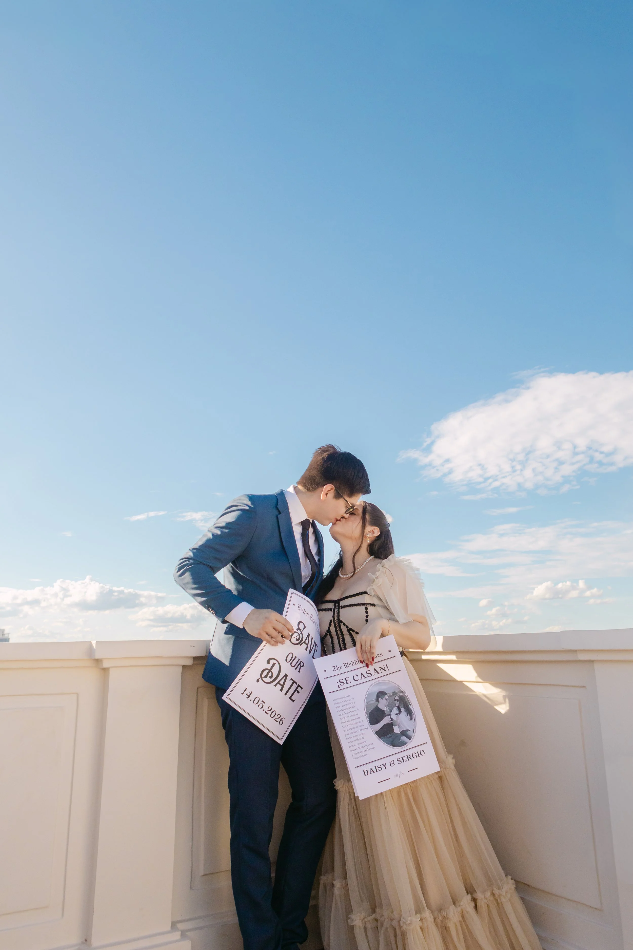 Una pareja se besa en un balcón, la mujer lleva vestido largo beige y el hombre traje azul, sosteniendo carteles de boda.