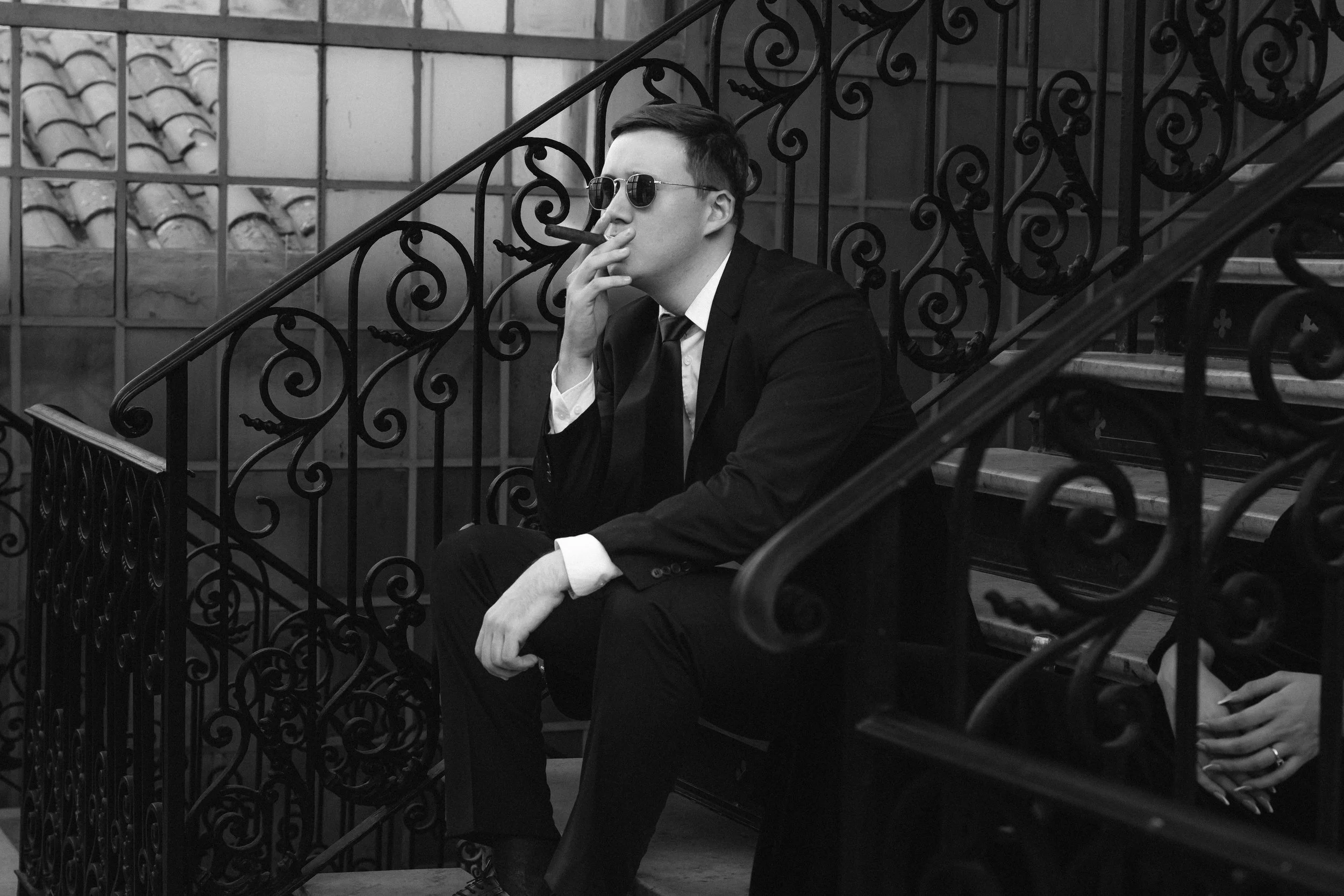 Un hombre vestido con traje negro, usando gafas oscuras, sentado en unas escaleras de hierro con barandilla decorativa, en un entorno que parece una terraza o balcón, en un ambiente urbano y elegante.