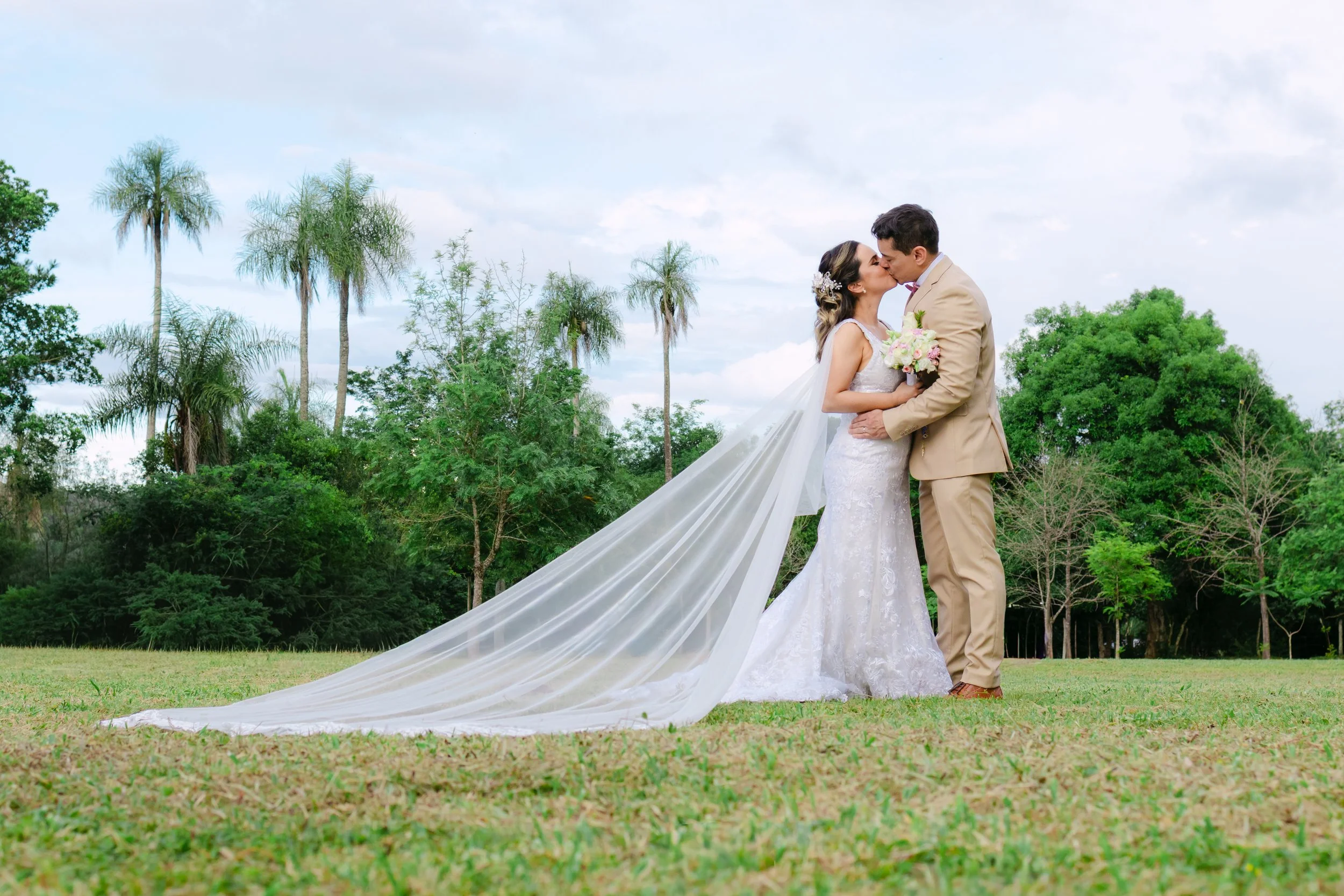 Pareja de novios besándose en un campo, con árboles y palmeras de fondo, ella con vestido de novia y él con traje beige, ella con velo largo y ramo de flores.
