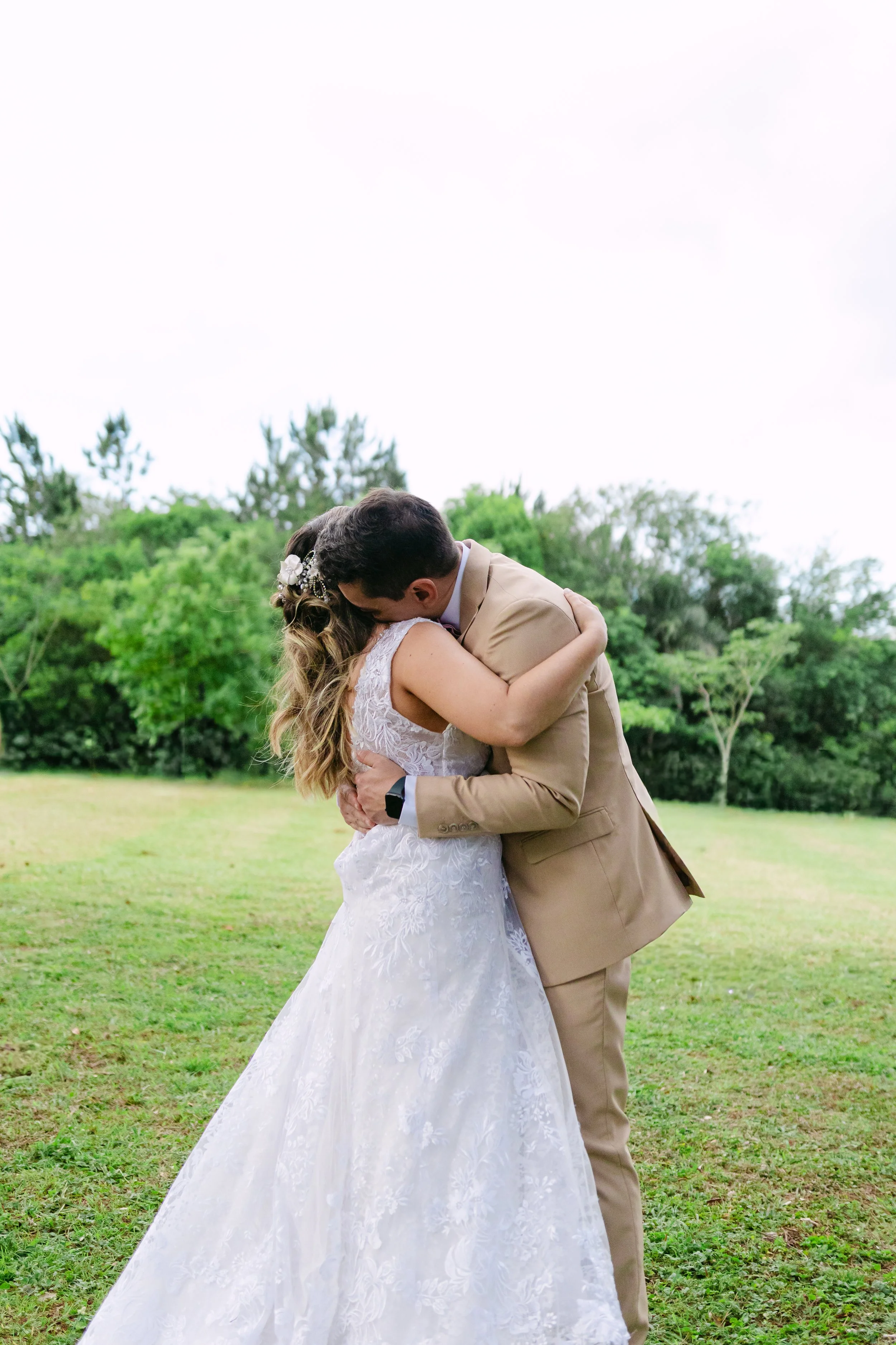 Una pareja de novios abrazándose en un campo, la novia viste un vestido de novia blanco con detalles de encaje y el novio lleva un traje beige.