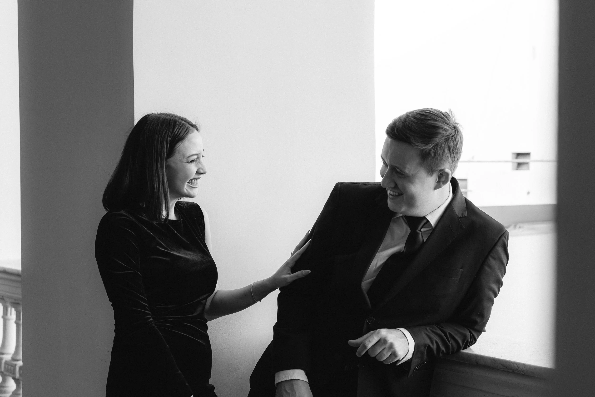Dos personas, un hombre y una mujer, riendo y conversando en un ambiente de oficina. La mujer lleva un vestido negro y el hombre un traje oscuro con corbata.