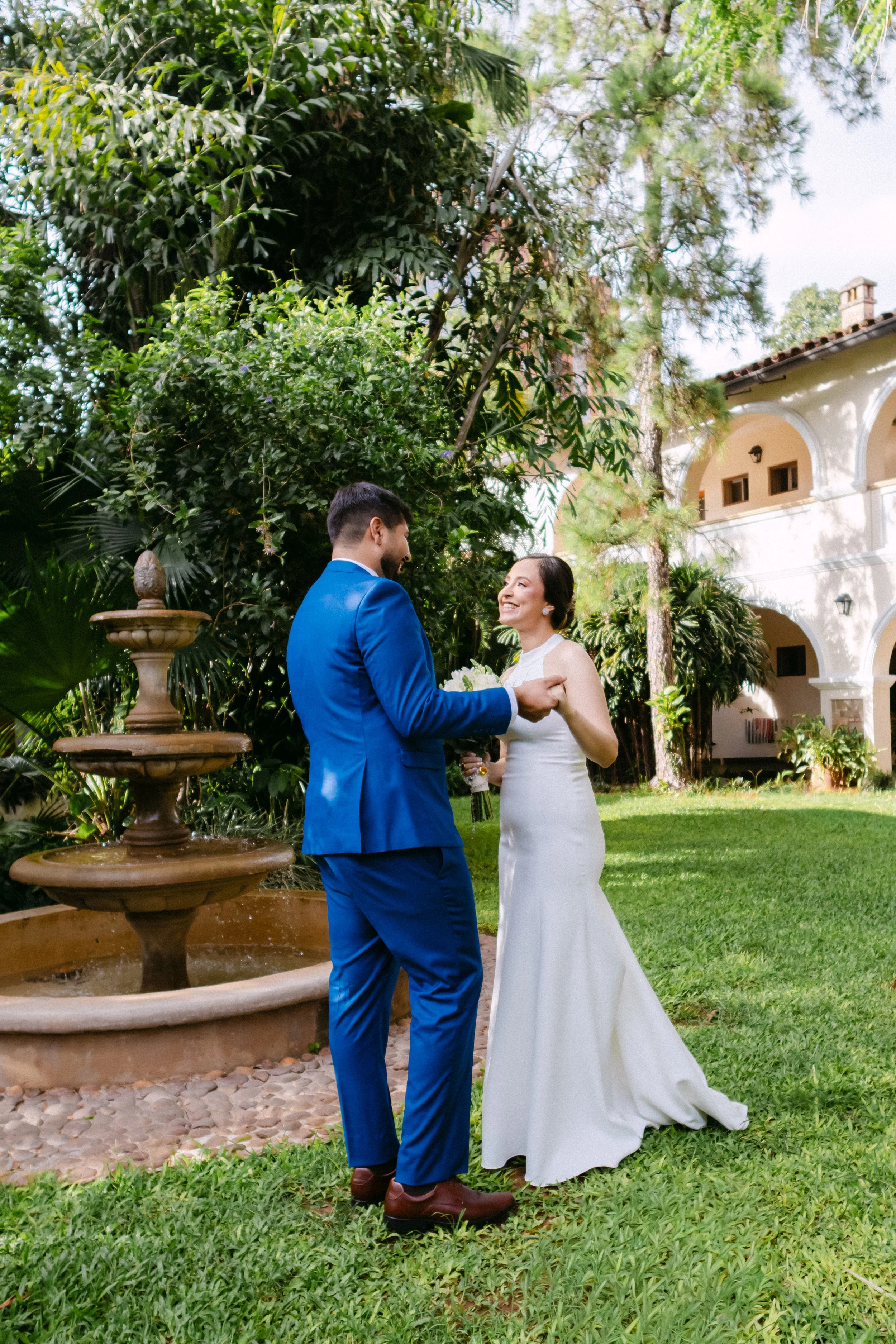 Una pareja de novios, la mujer con vestido blanco y el hombre con traje azul, se miran y participan en una ceremonia en un jardín con fuente y árboles.