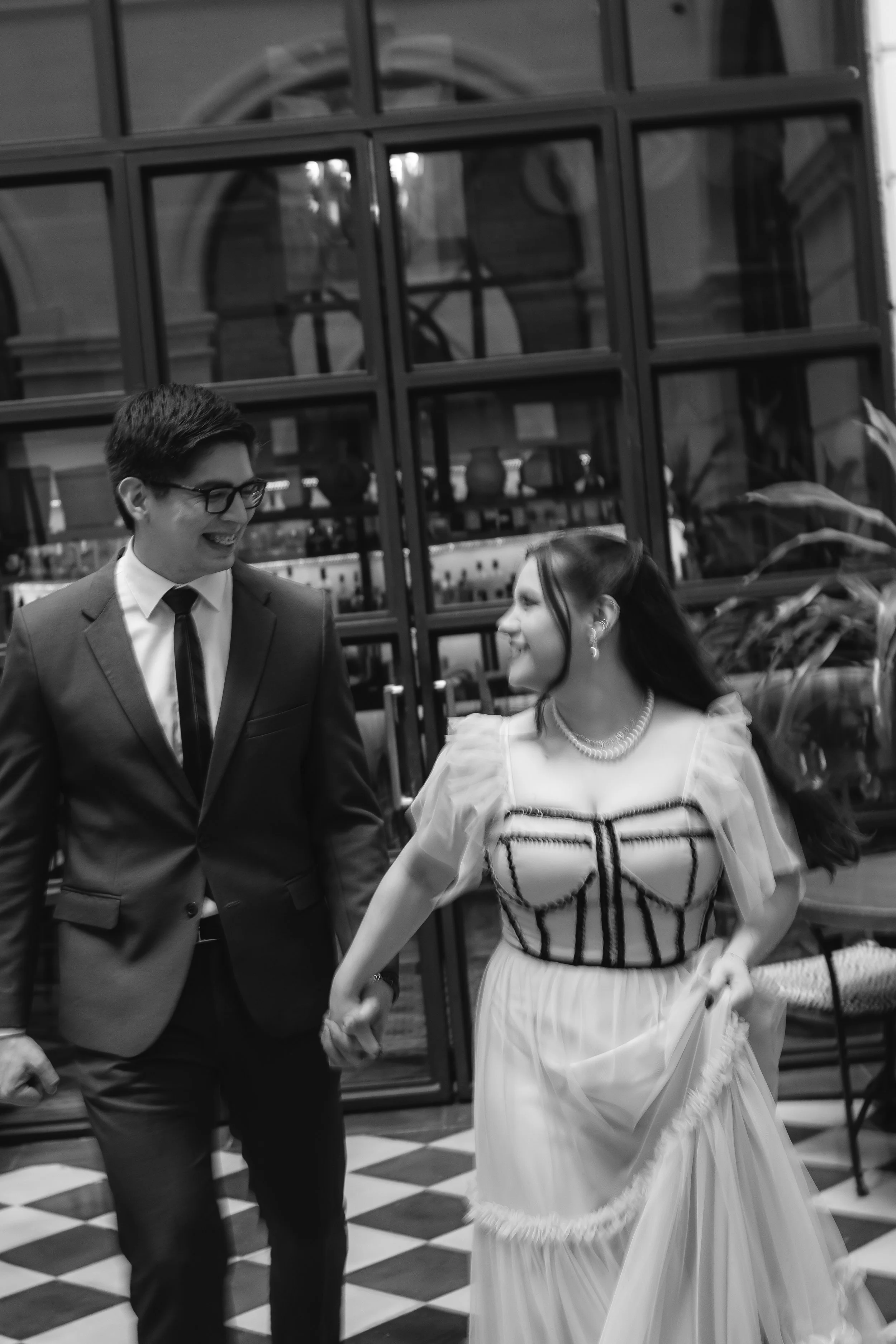 Una pareja bailando y sonriendo, en un lugar con ventanas de vidrio y piso de cuadros, en blanco y negro.