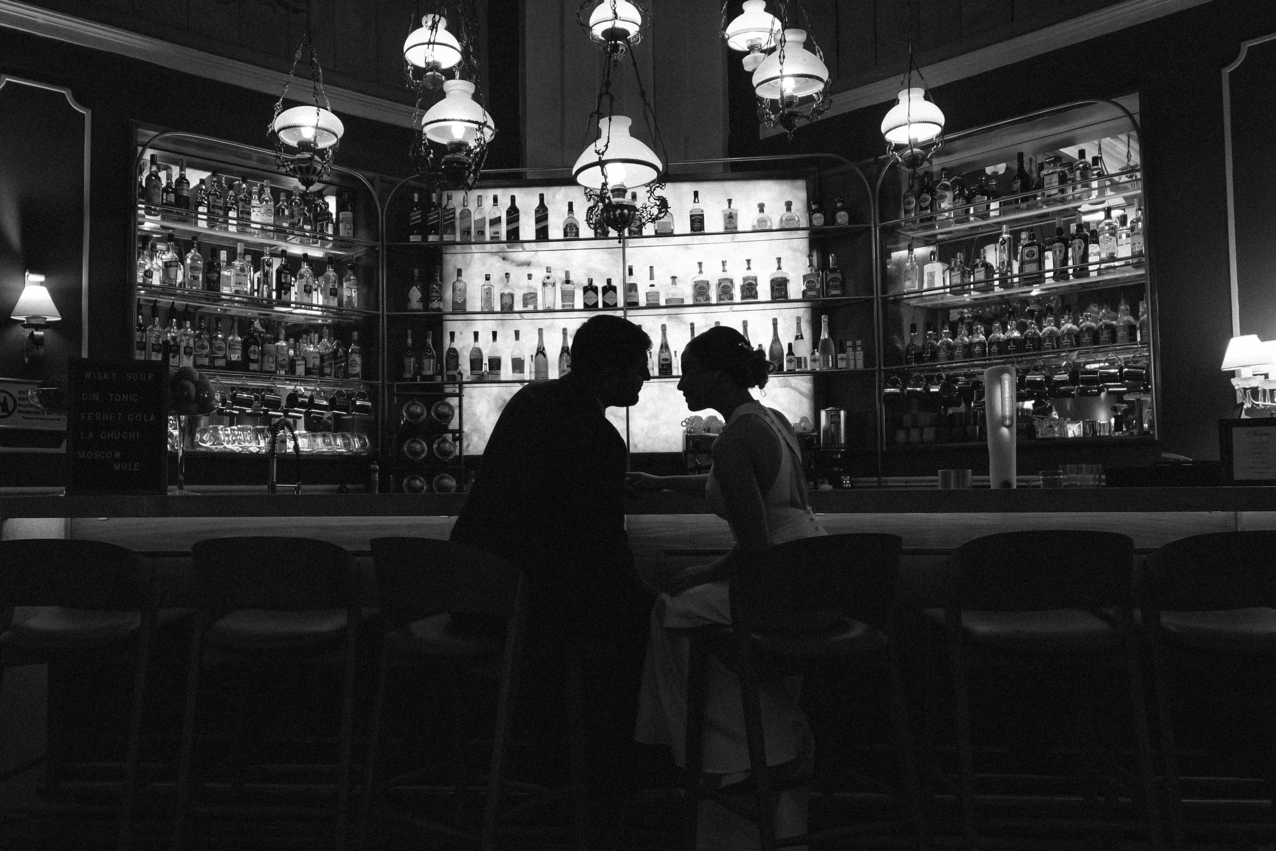 Pareja en un bar, vista en silueta, con estantes de botellas y lámparas en el fondo.