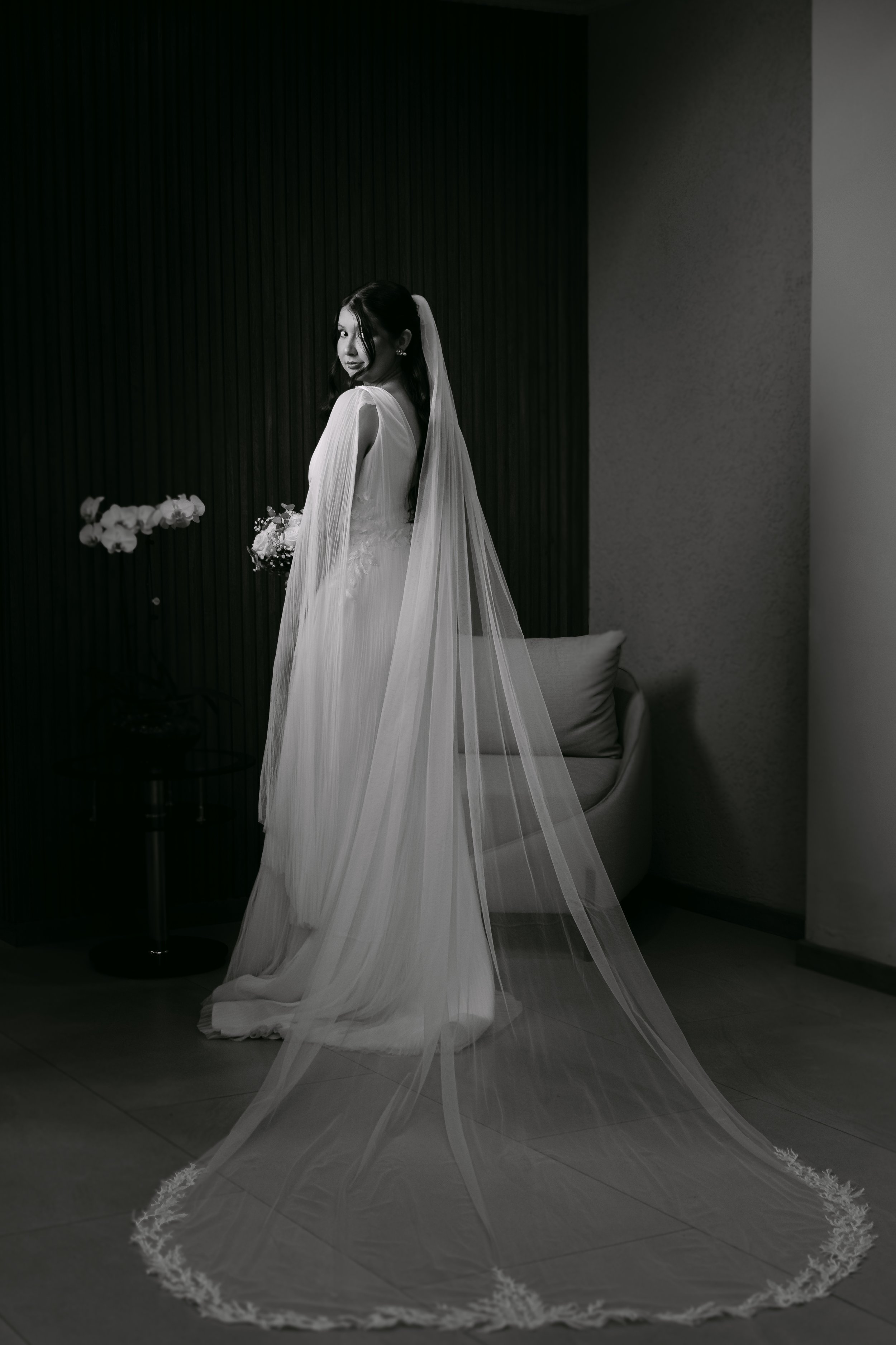 Mujer vestida de novia con velo largo en interior con sillas y flores.