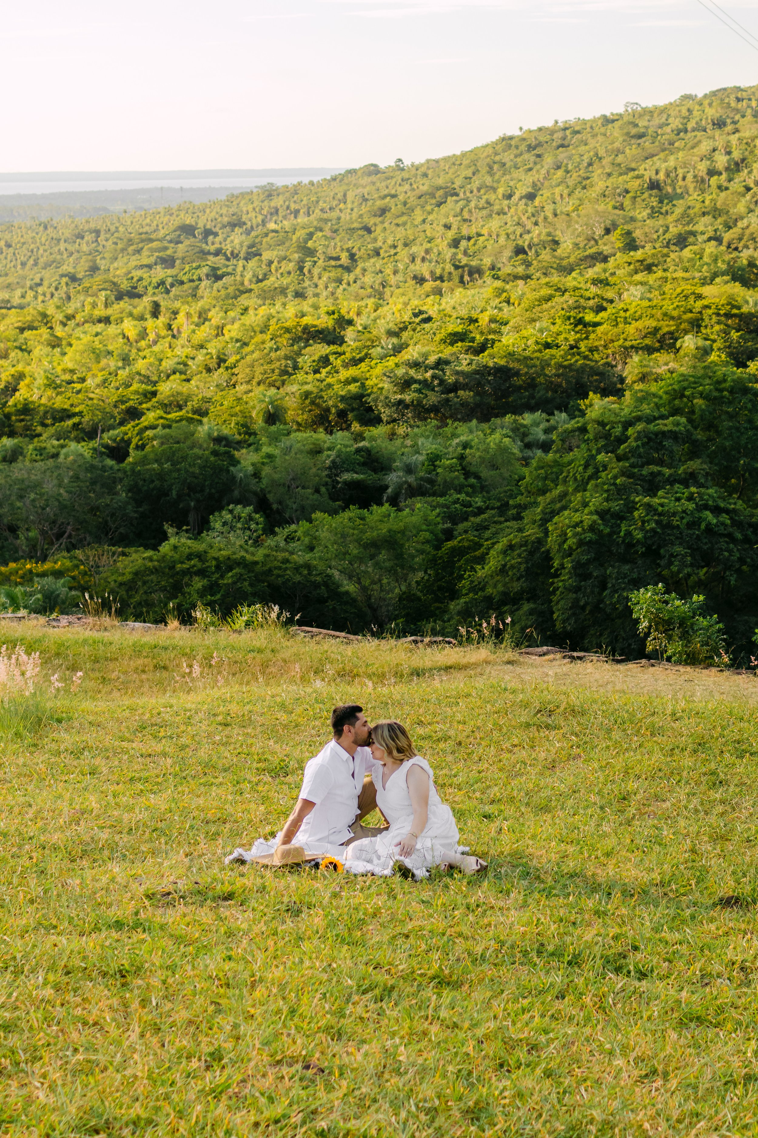 Una pareja sentada en el césped en un paisaje natural con árboles y montañas al fondo, en un día soleado.