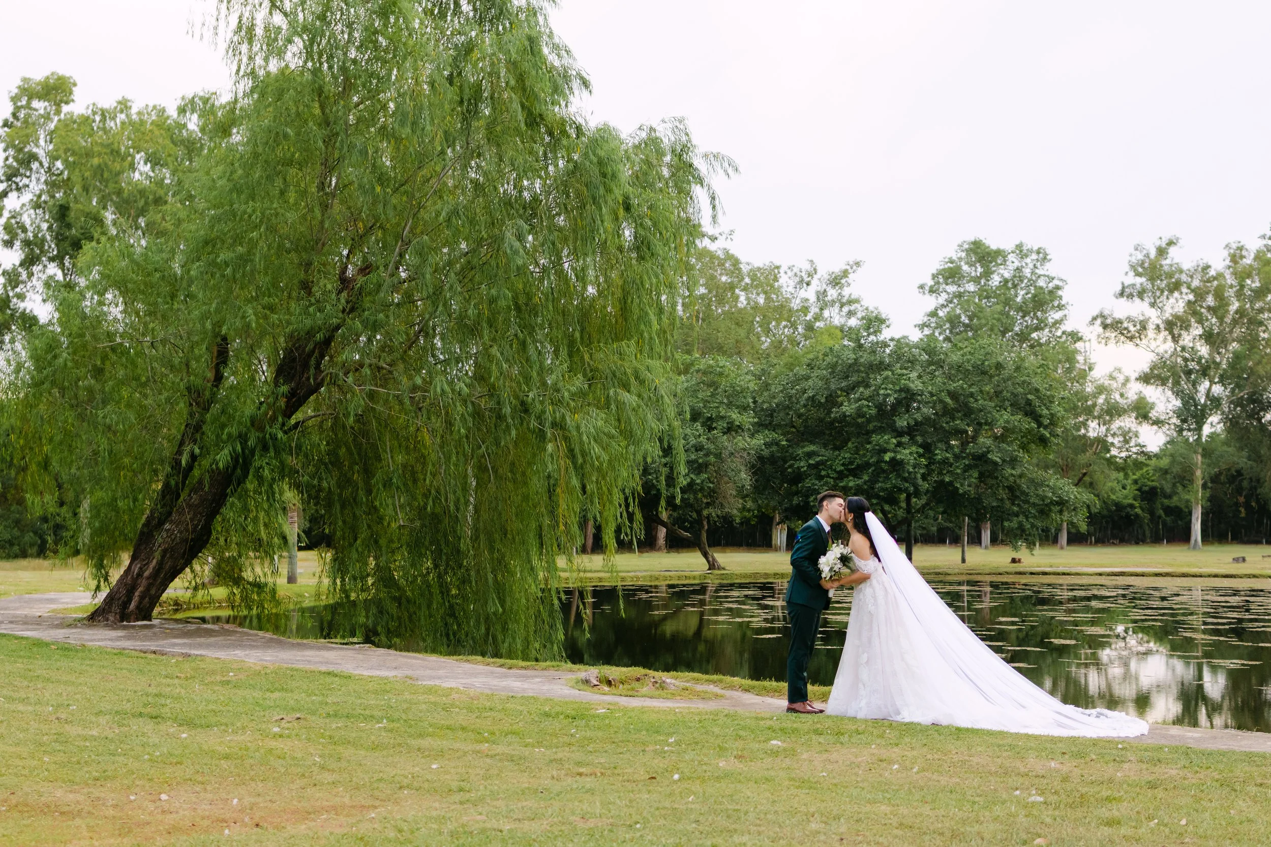 Una pareja de recién casados besándose junto a un estanque en un parque rodeado de árboles.