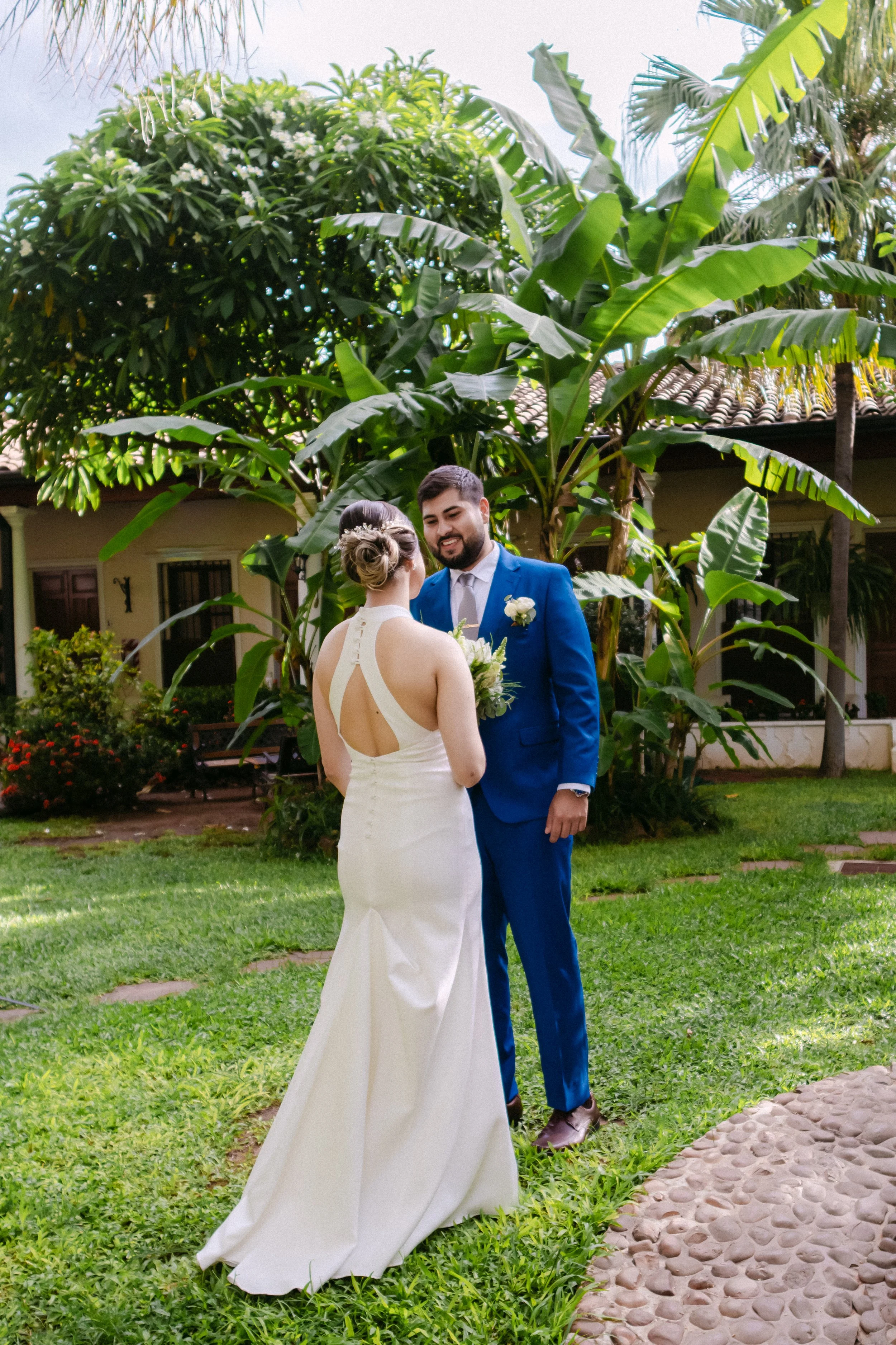 Pareja de novios en una boda al aire libre, con vegetación y plantas grandes en el fondo, el hombre viste traje azul y la mujer vestido blanco, ella sostiene un ramo de flores.