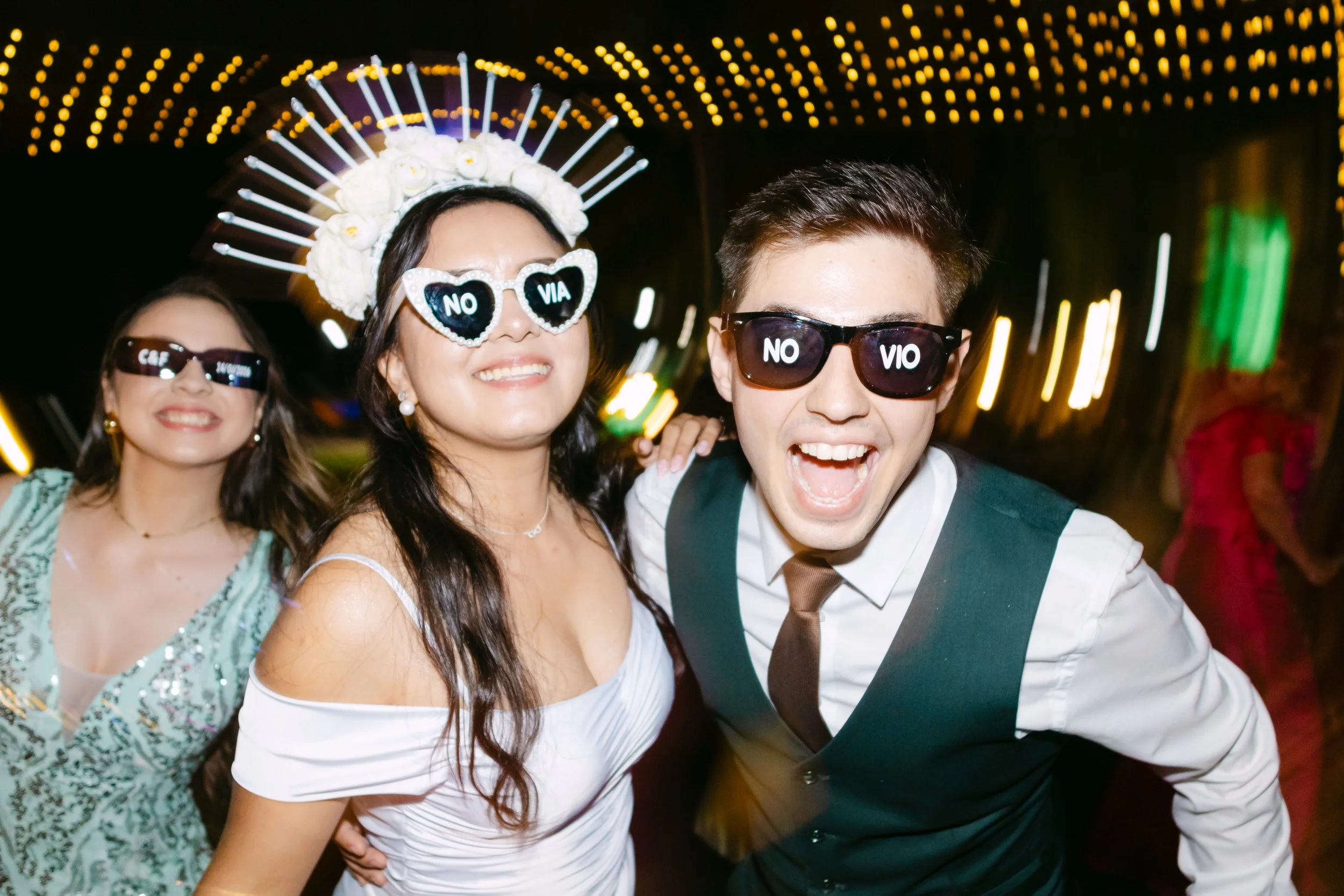Tres personas en una fiesta, con gafas que dicen 'No Vio', una mujer con corona y vestido blanco, un hombre con gafas y corbata, otra mujer con gafas y vestido verde y un fondo de luces coloridas