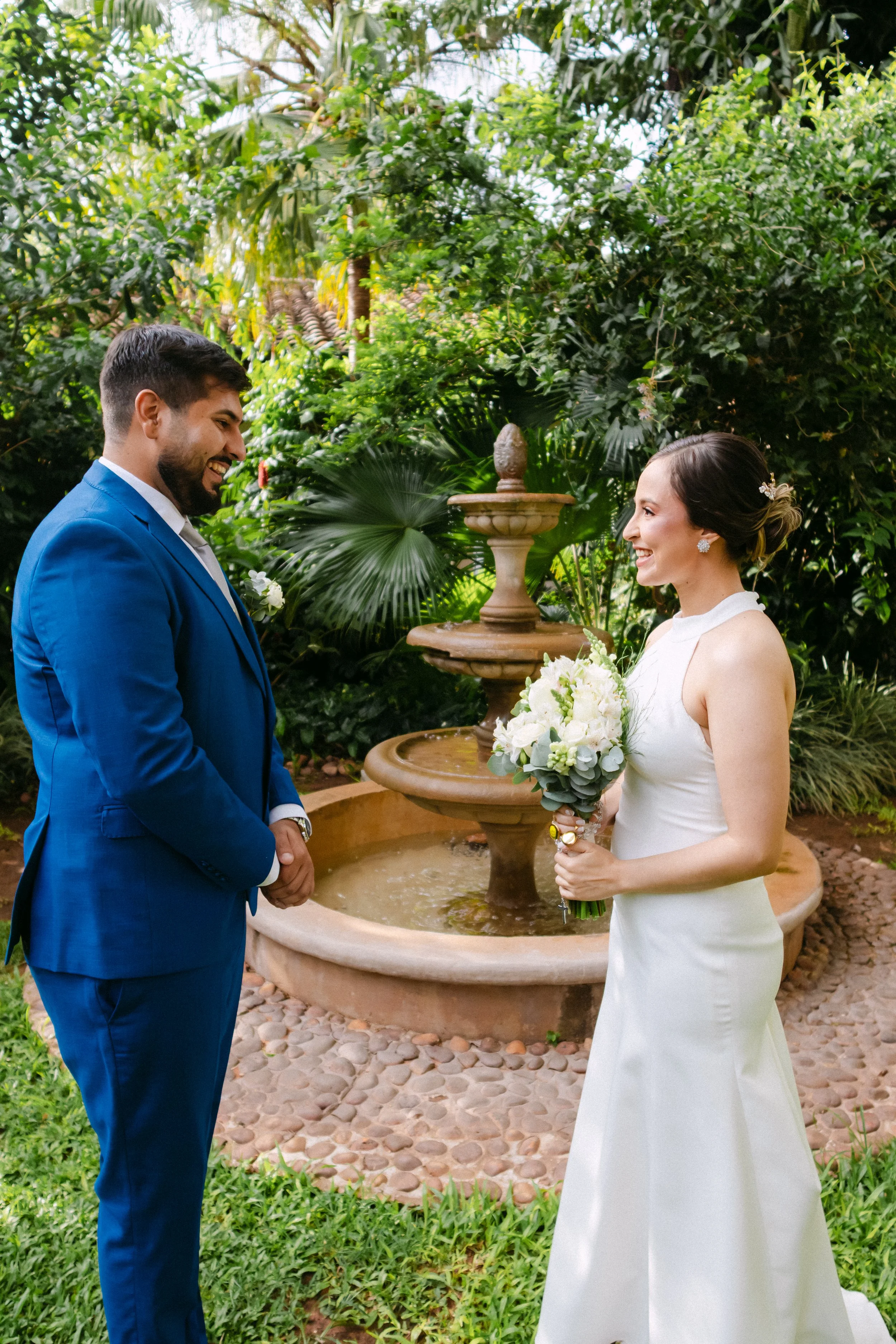 Una pareja en boda en un jardín, la mujer con vestido blanco y bouquet, el hombre con traje azul, frente a una fuente de agua