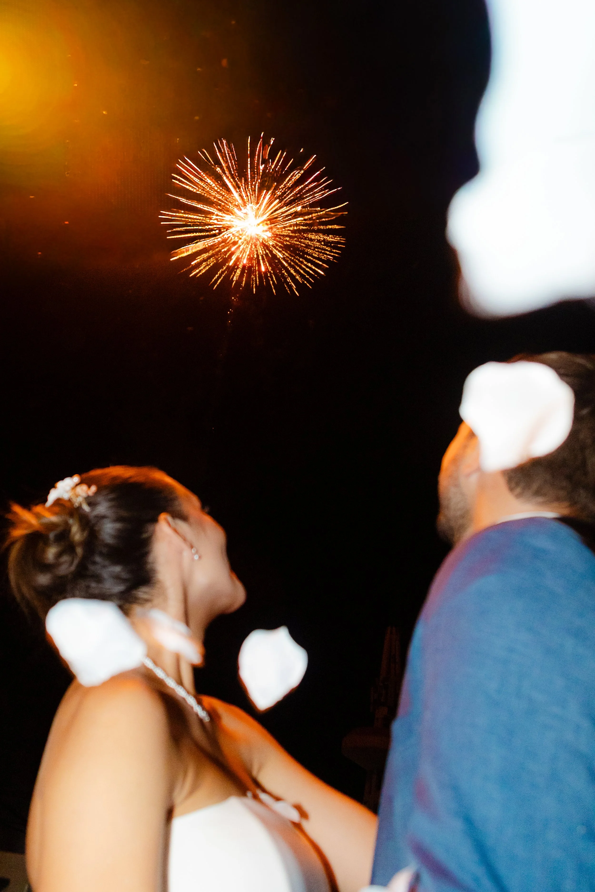 Una pareja viendo fuegos artificiales en la noche. La mujer lleva un vestido blanco y un peinado con recogido. El hombre viste una chaqueta azul y barba. Hay pétalos blancos en el aire y un cielo oscuro con fuegos artificiales de color dorado en el fondo.
