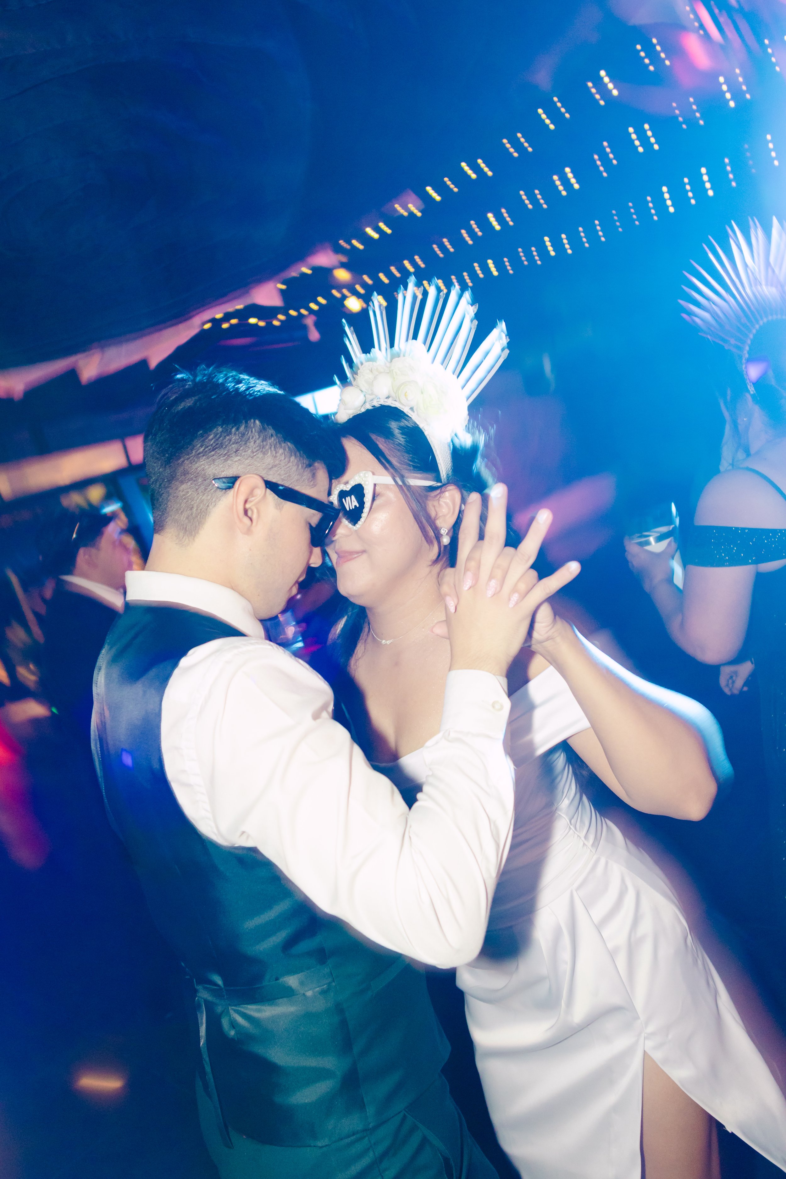 Pareja bailando en una fiesta, la mujer lleva una corona de flores y gafas de sol blancas, el hombre viste un traje formal, ambiente con luces y decoraciones vibrantes.