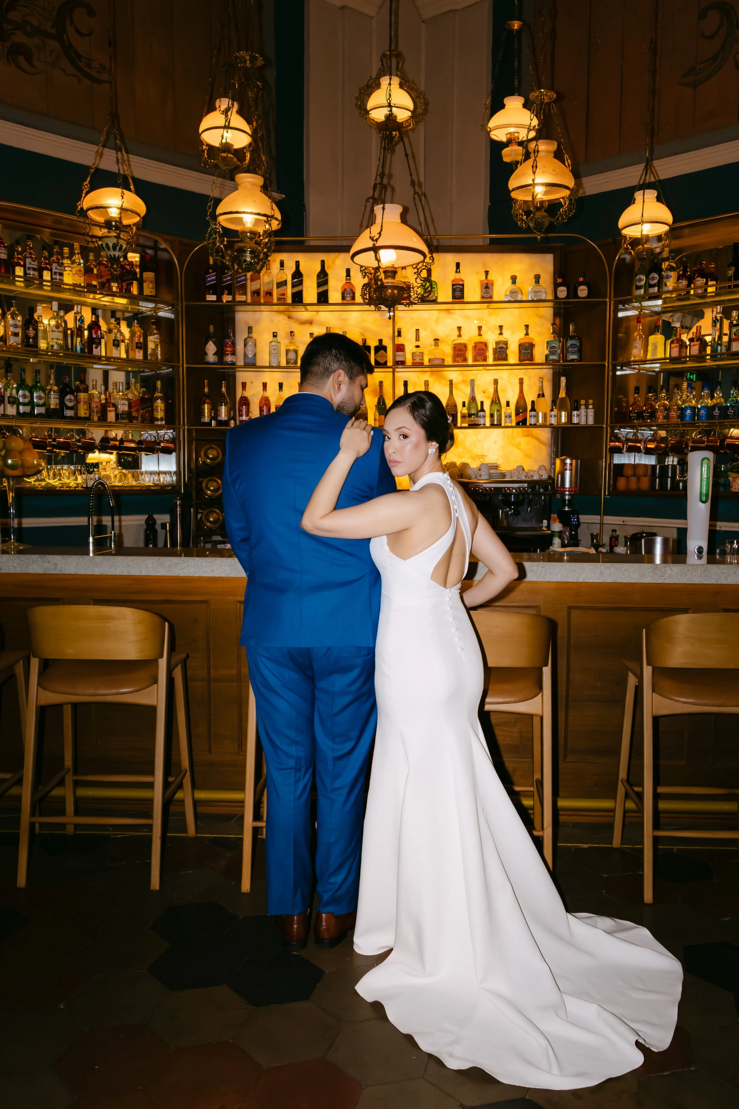 Pareja de novios en un bar elegante, la mujer con vestido de novia y el hombre con esmoquin azul, frente a una barra de bar con muchas botellas de alcohol y lámparas colgantes.