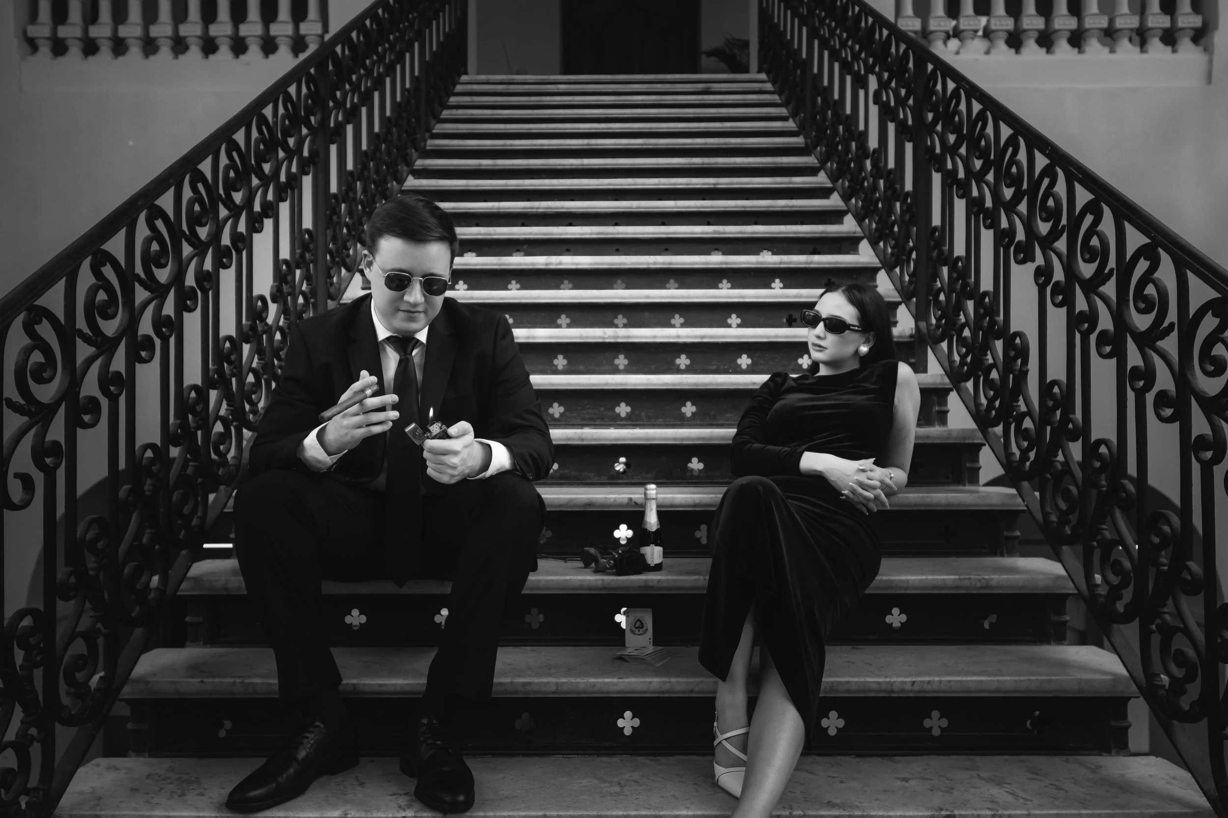 Dos personas vestidas con ropa elegante, un hombre y una mujer, sentados en unas escaleras de hierro forjado. El hombre lleva traje negro, camisa blanca y gafas de sol, y está usando un cigarro y un teléfono móvil. La mujer lleva un vestido largo y t