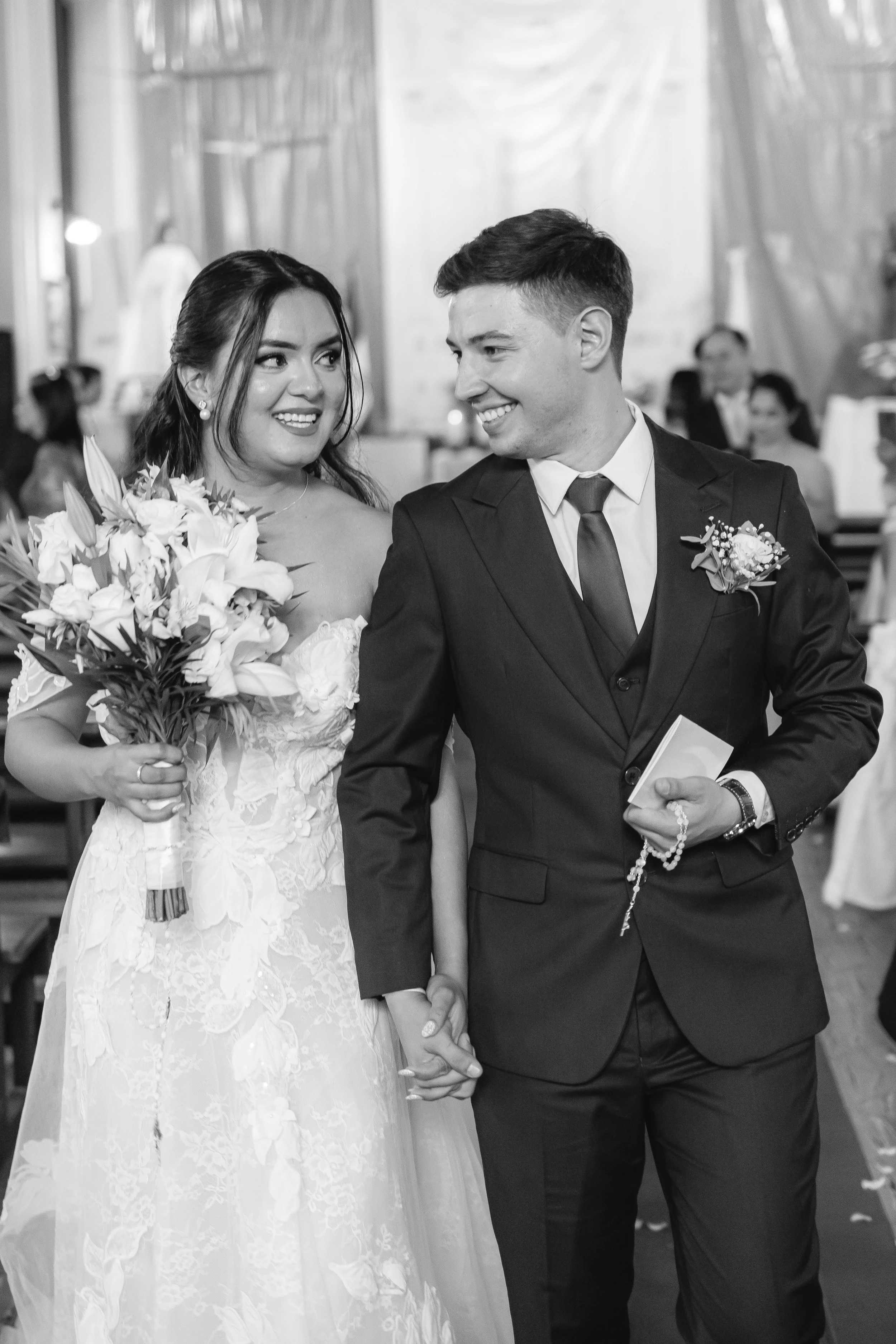 Una pareja de novios en su boda, la novia sostiene un ramo de flores y mira con cariño al novio, que viste un traje oscuro y sostiene un rosario.