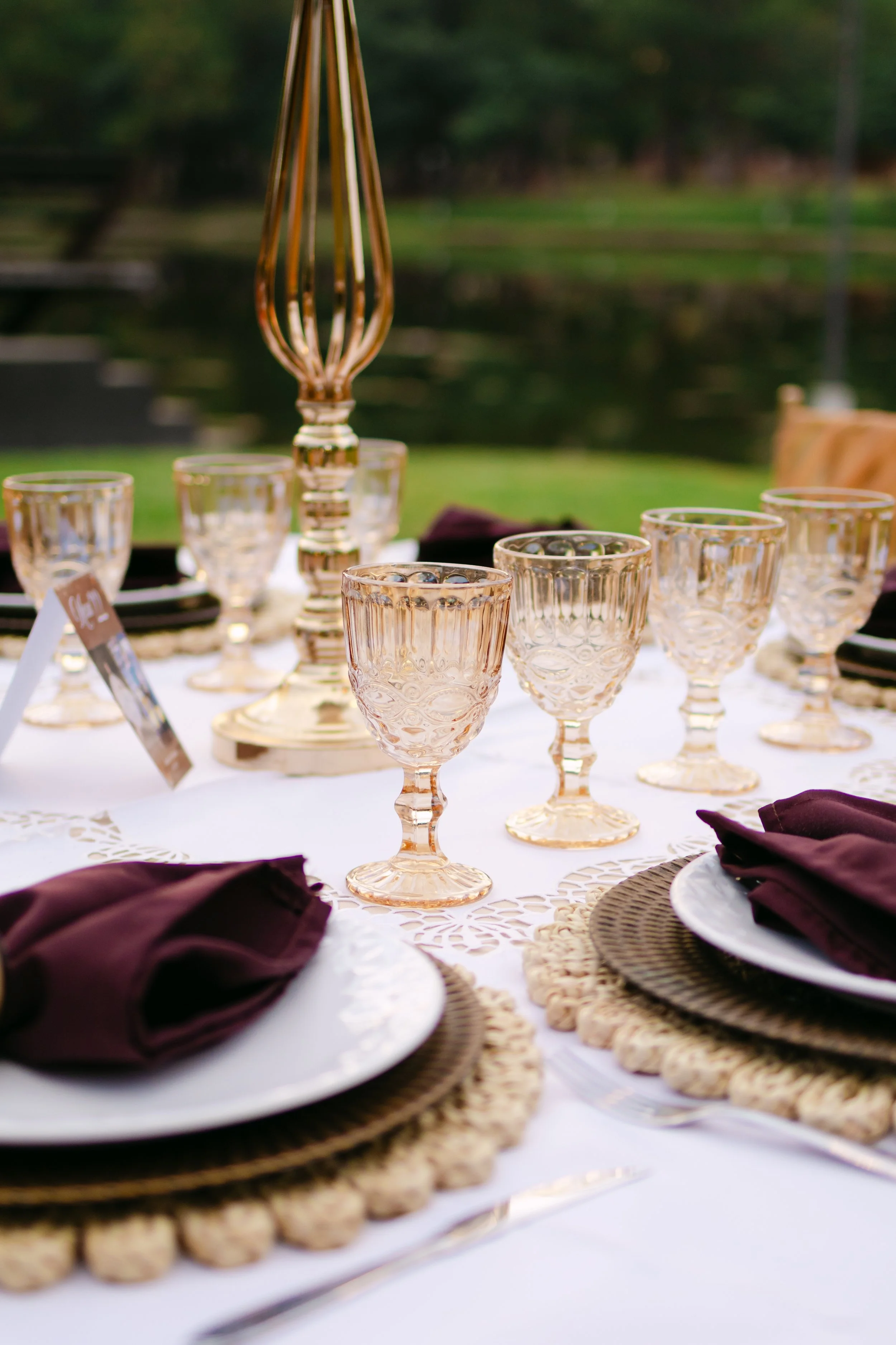 Mesa decorada con cristalería, platos, tenedores, servilletas moradas y un centro de mesa con un jarrón decorativo, en un entorno al aire libre con fondo de árboles y un lago.