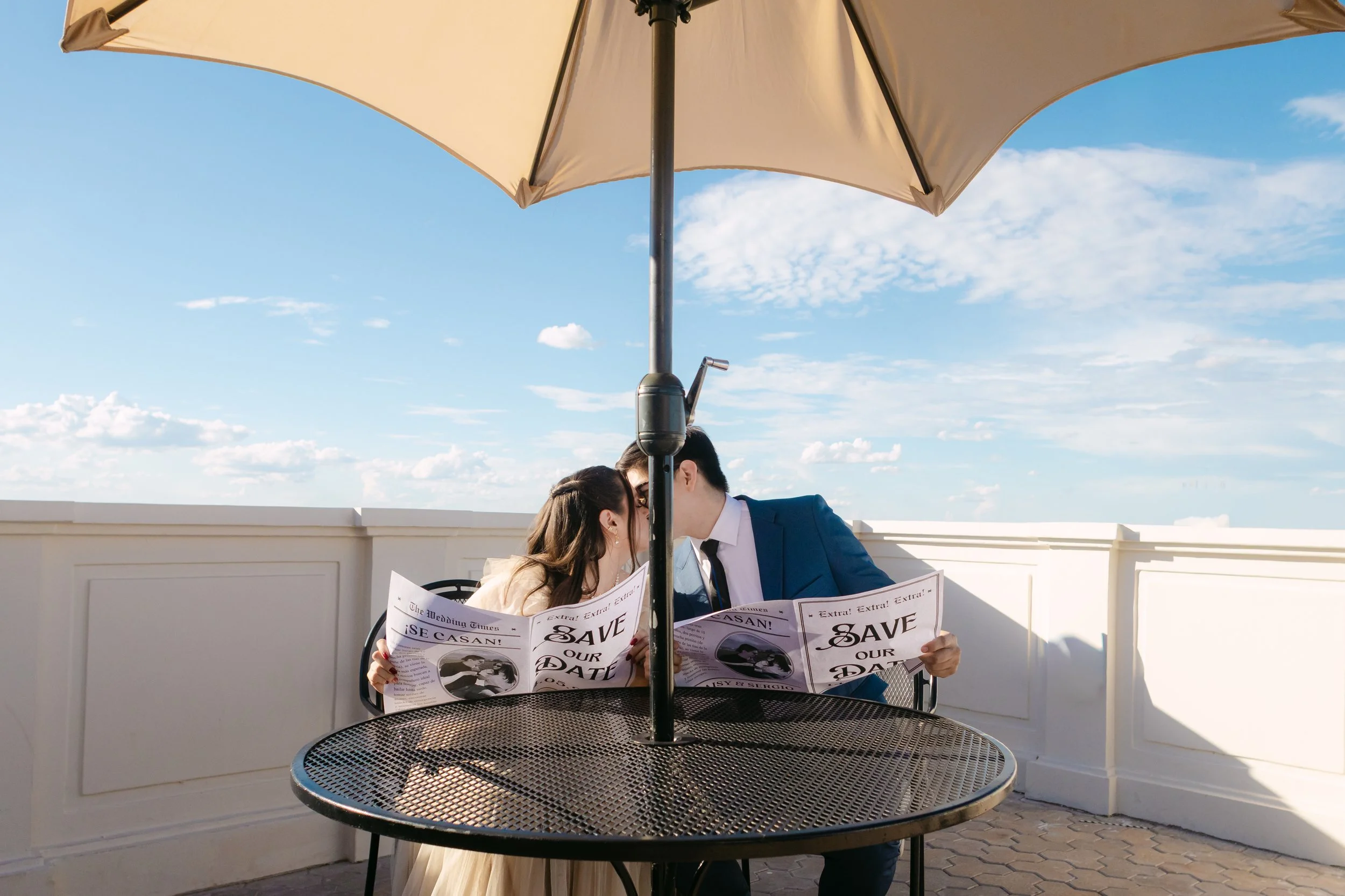 Una pareja joven en traje formal sentados en una mesa de hierro en una terraza con un paraguas grande, leyendo periódicos y tocándose con la frente, en un día soleado con cielo despejado.