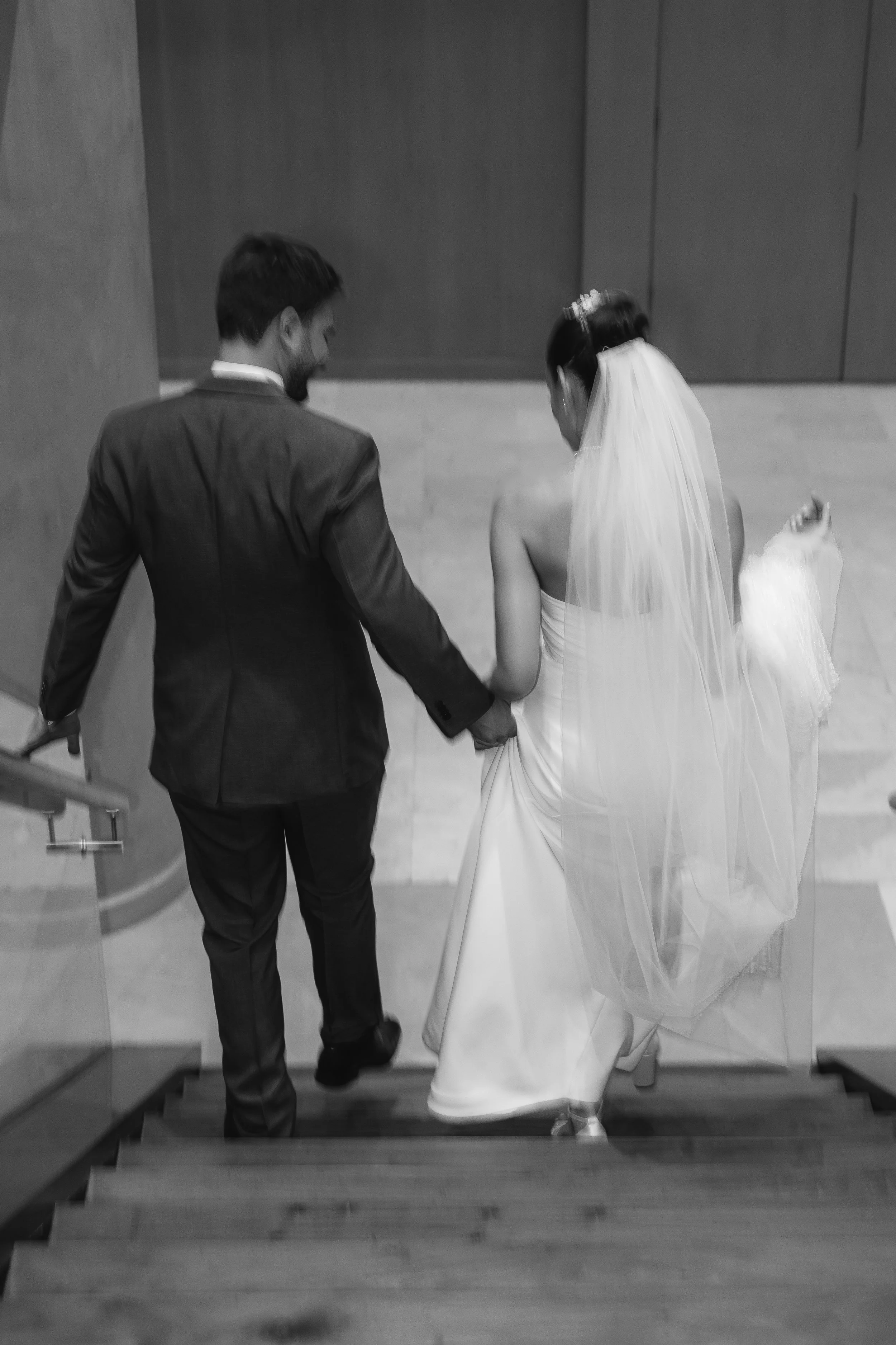 Pareja de novios bajando una escalera, ella con vestido de novia y velo, él con traje formal, en un ambiente interior.