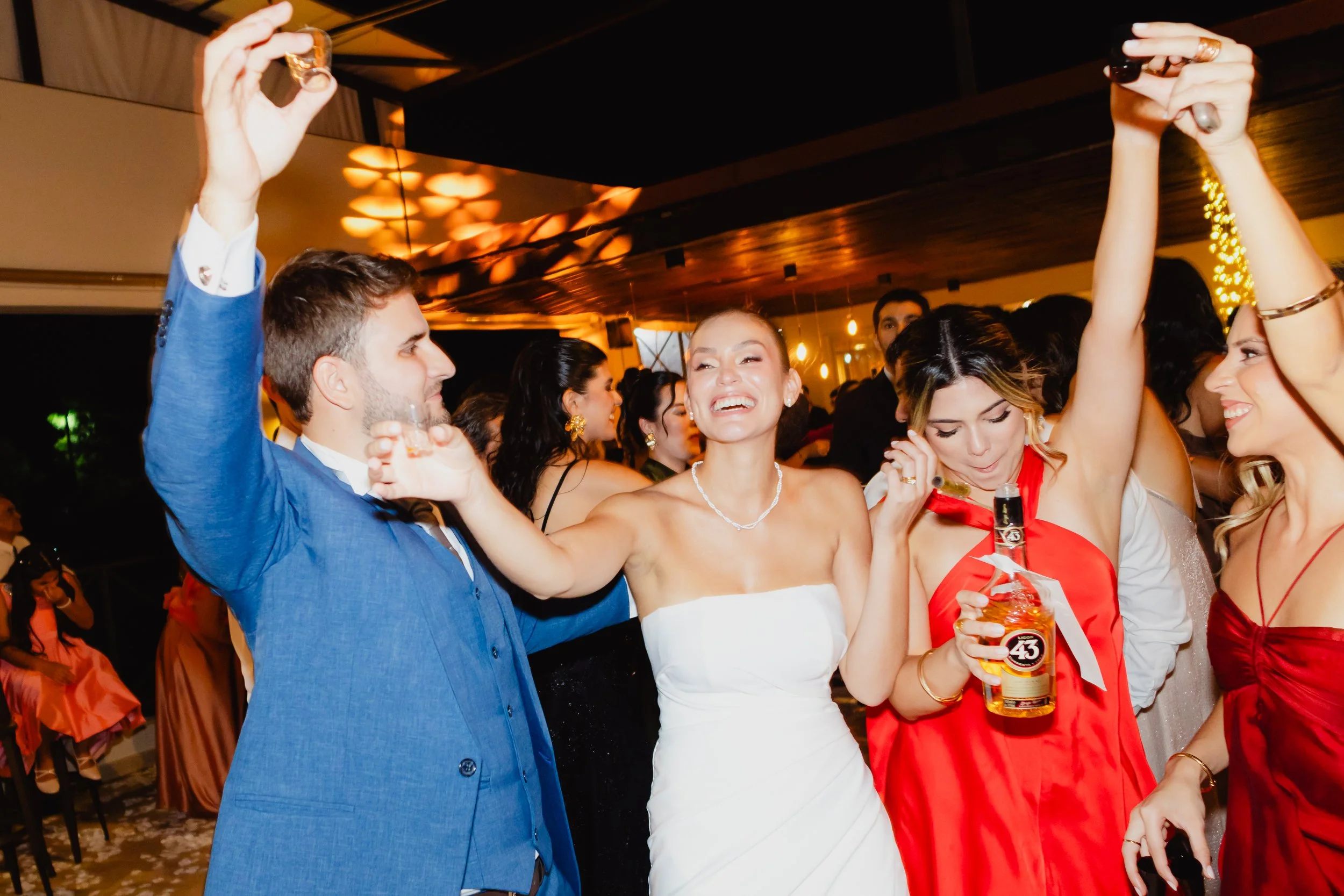 Grupo de personas bailando y disfrutando en una fiesta o celebración en interiores, rodeados de luces cálidas y decoraciones festivas.