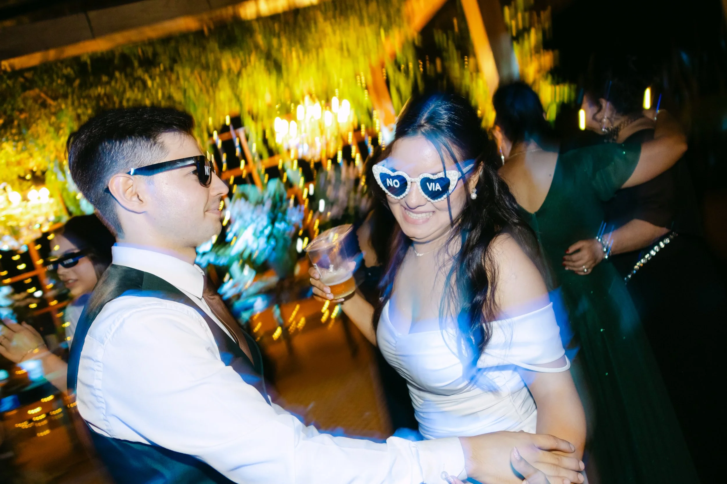 Pareja disfrutando en una fiesta, la mujer con gafas que dicen 'No Via', el hombre con gafas oscuras, ambos bailando y sonriendo en un ambiente festivo con luces y otras personas al fondo.
