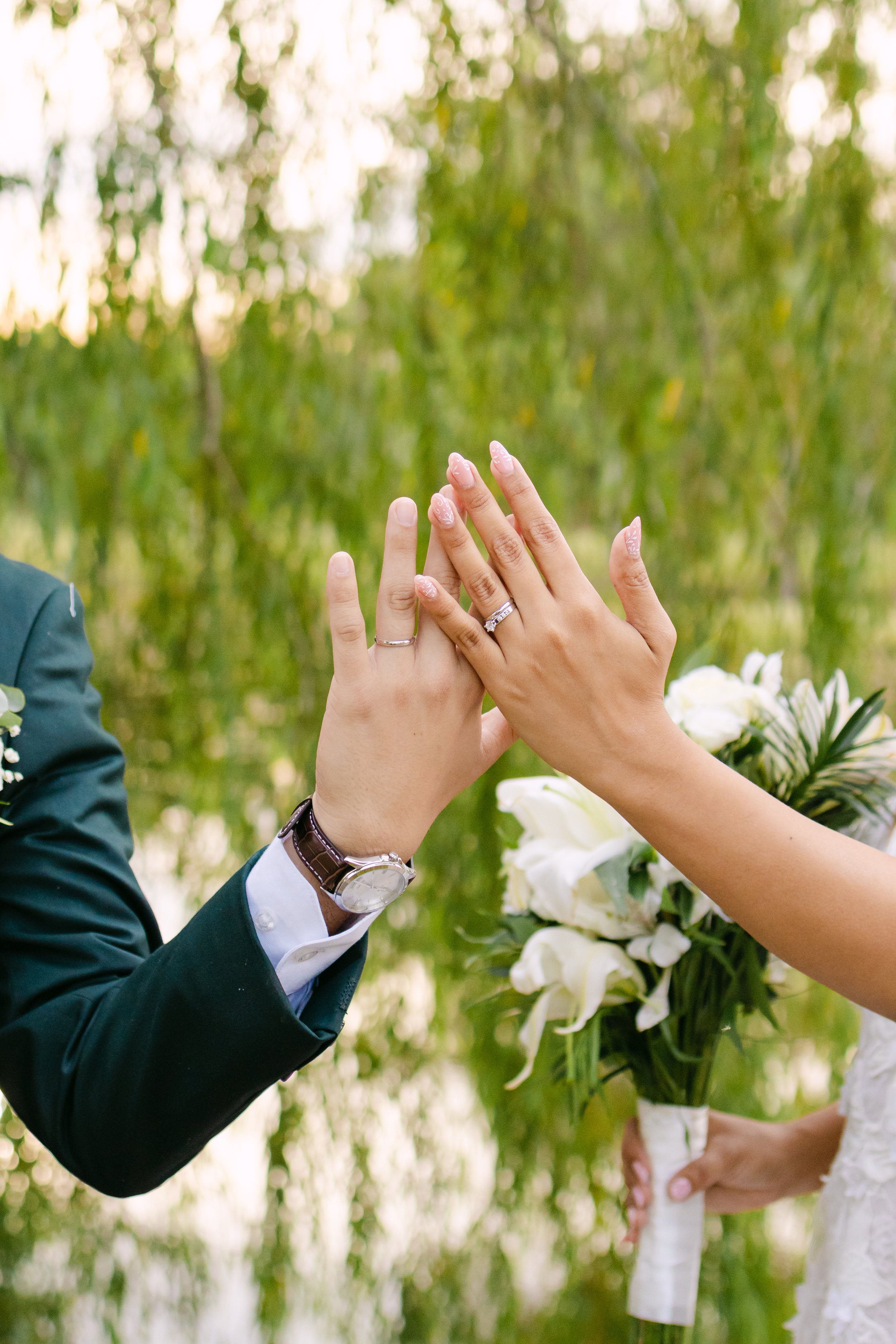 Mano y mujer con anillos de boda tocándose en señal de unión, en un entorno natural con flores blancas y fondo de árboles verdes.