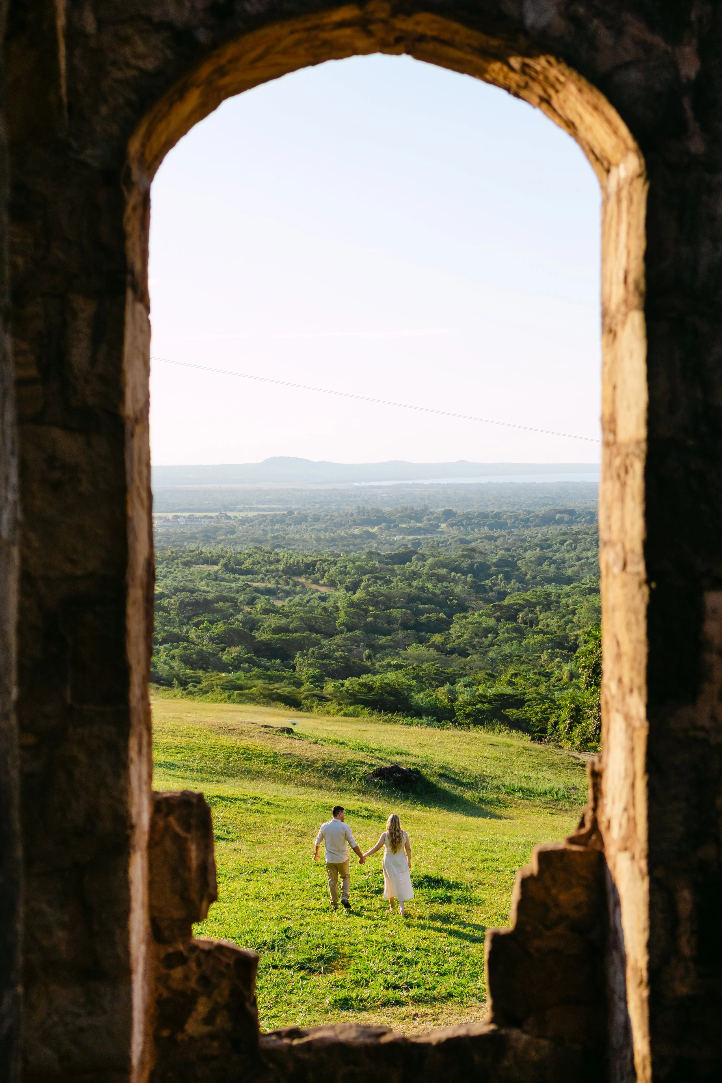 Una pareja caminando de la mano en un campo verde, vista a través de una ventana de piedra con arco, con un paisaje de colinas y árboles al fondo.