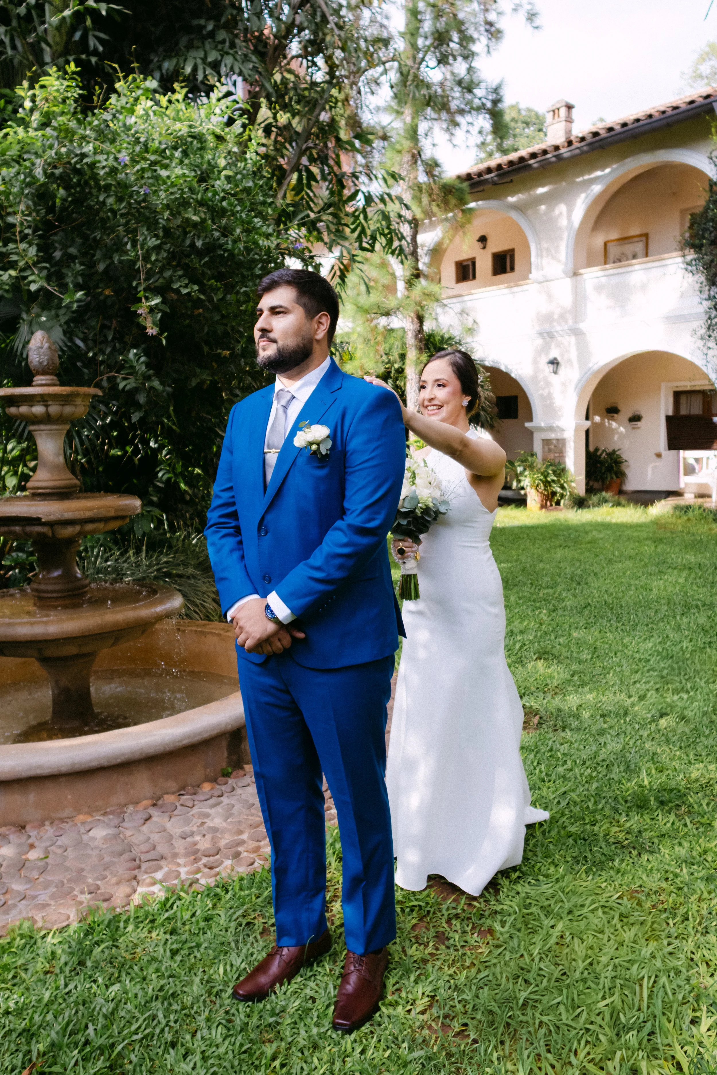 Una pareja en una boda, la mujer está sonriendo y colocando la mano en el hombro del hombre, quien viste un traje azul. Están en un jardín con una fuente y una casa colonial en el fondo.