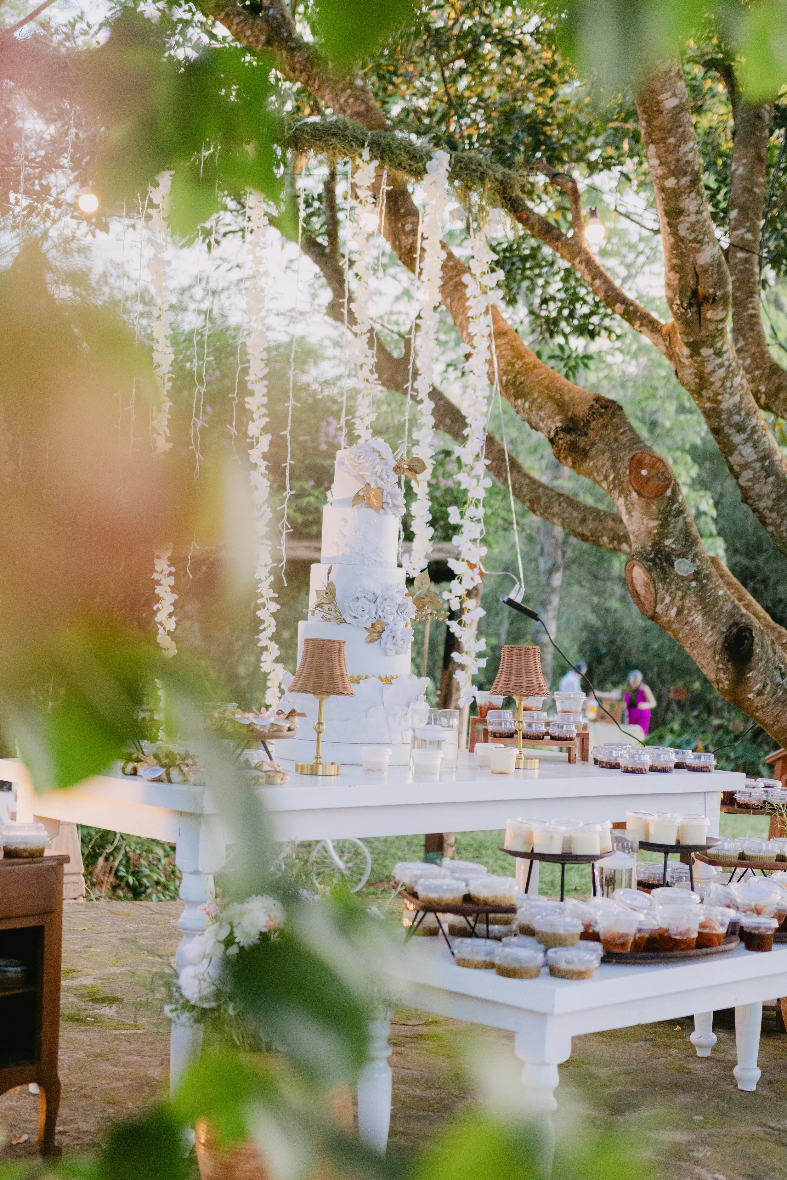 Mesa decorada para una celebración al aire libre con un pastel blanco de varios niveles, adornado con flores y detalles dorados, rodeada de dulces en frascos y lámparas pequeñas, en un entorno natural con árboles y luces.