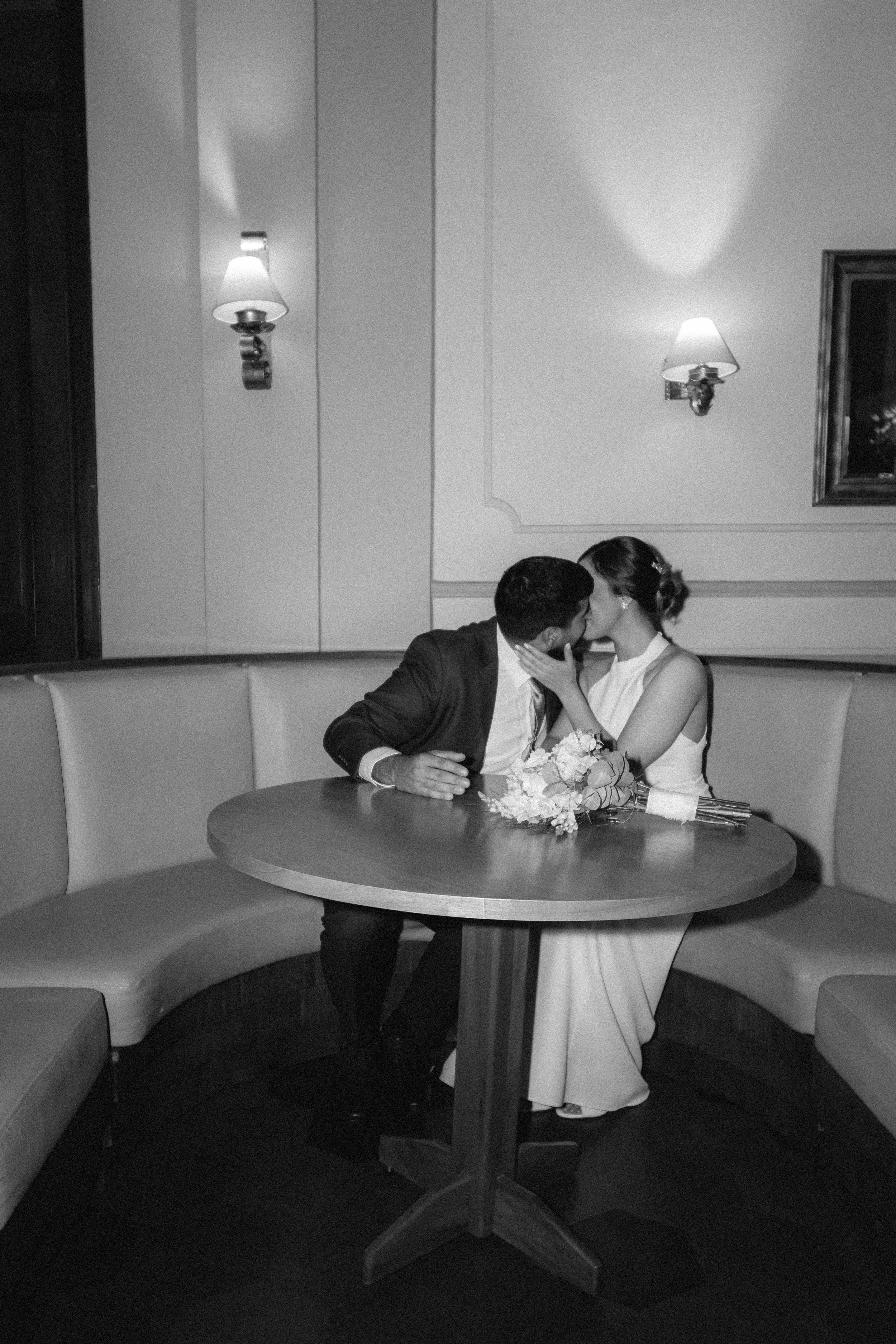 Una pareja se besa en una mesa en un restaurante elegante, con luces suaves y decoraciones sencillas.