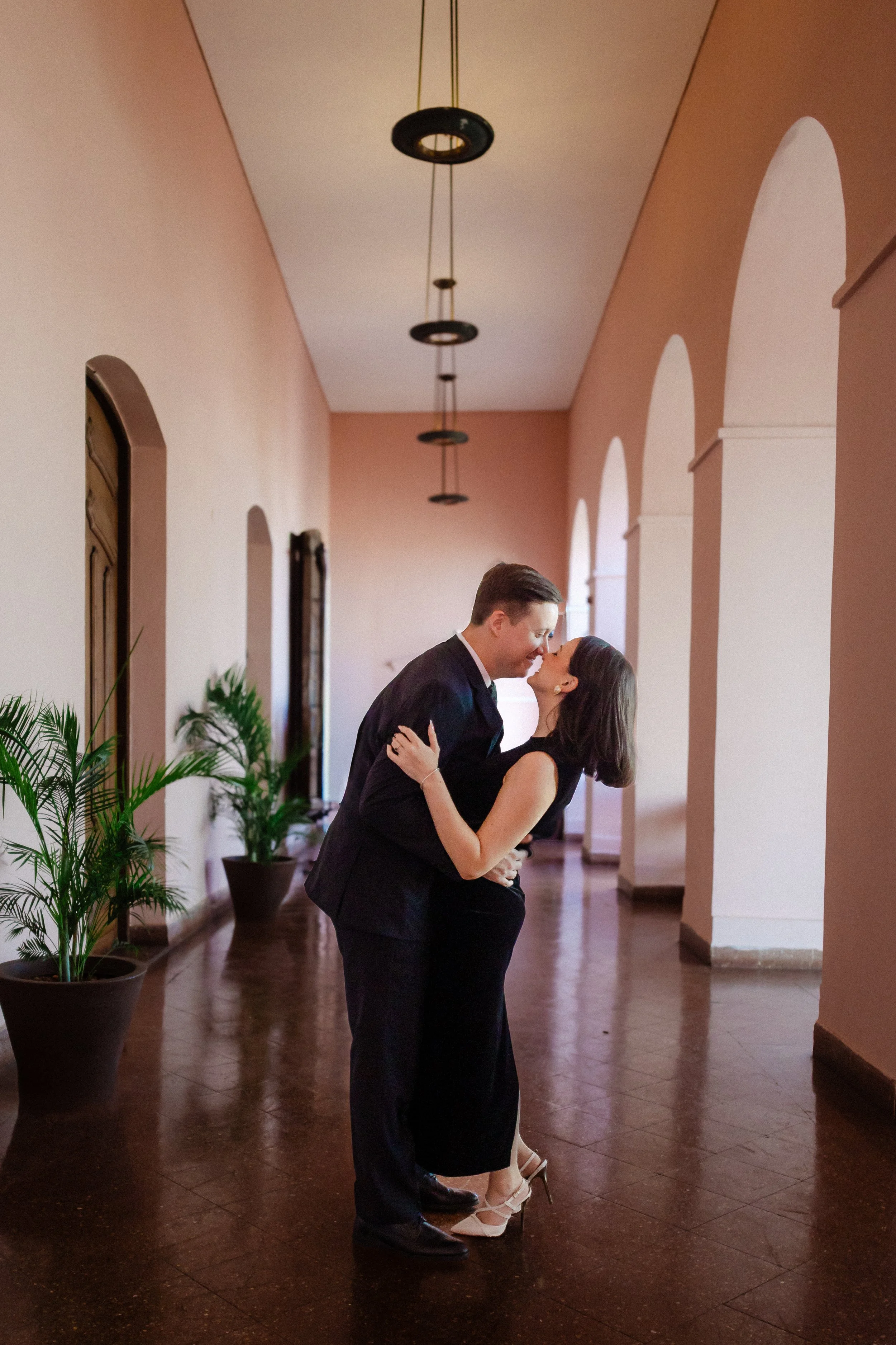 Pareja besándose en un pasillo con paredes color rosa, con plantas en macetas y ventanas arqueadas.