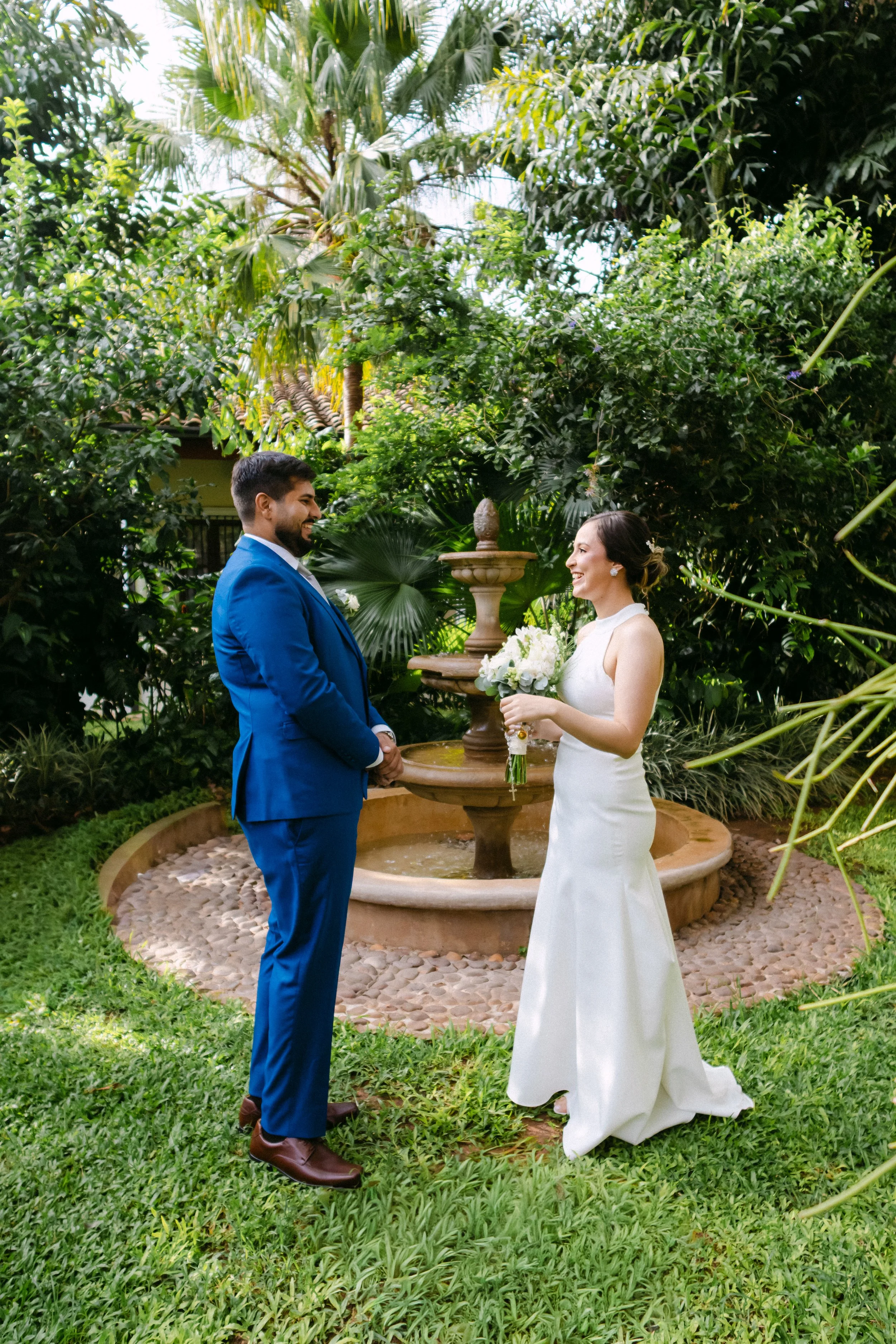 Pareja de recién casados en una boda al aire libre, la mujer con vestido de novia y el hombre con traje azul, en un jardín rodeado de vegetación y una fuente en el fondo.