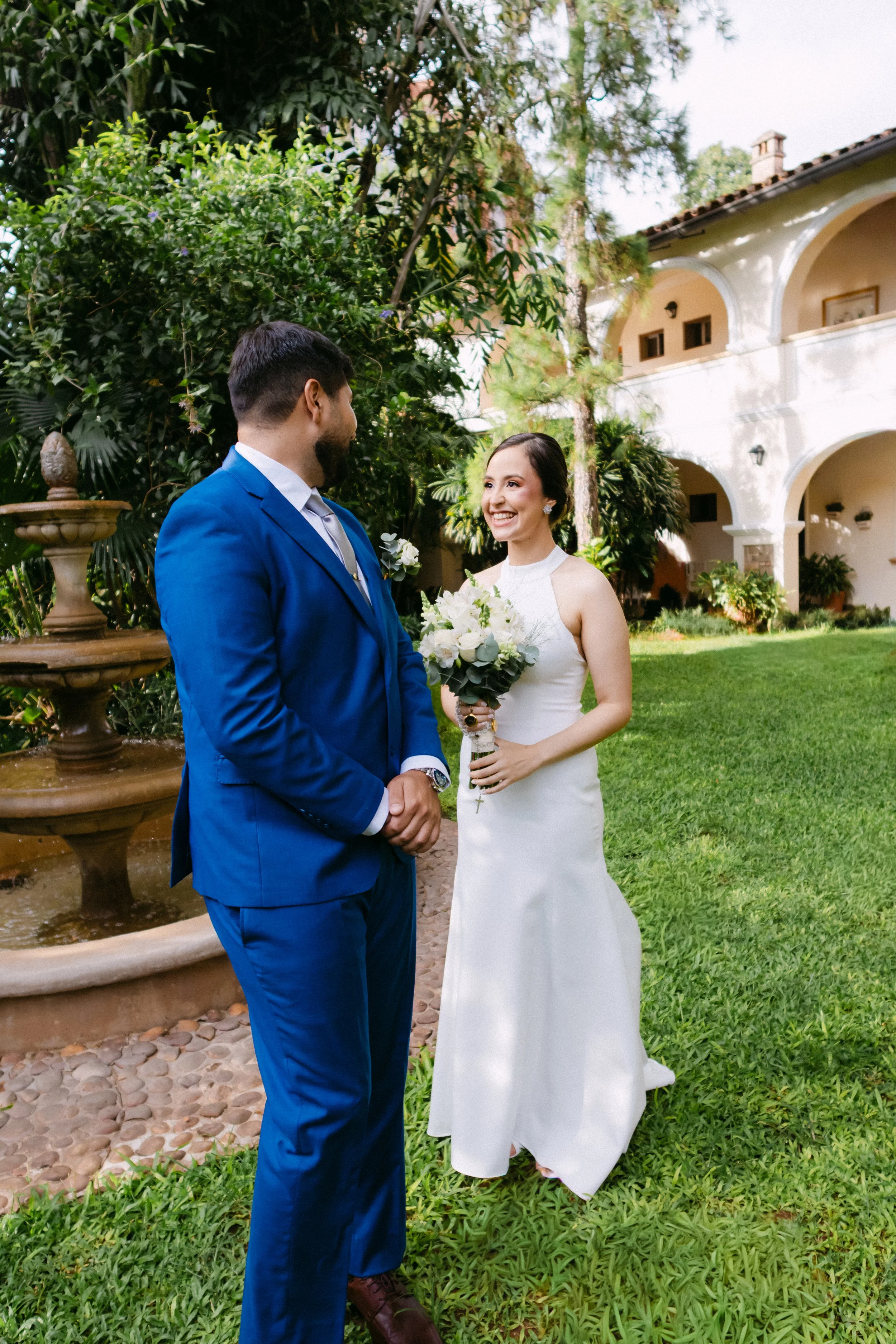 Pareja de novios en boda al aire libre, la novia sostiene un ramo de flores blancas y la ceremonia está en un jardín con fuente y casa colonial en el fondo.