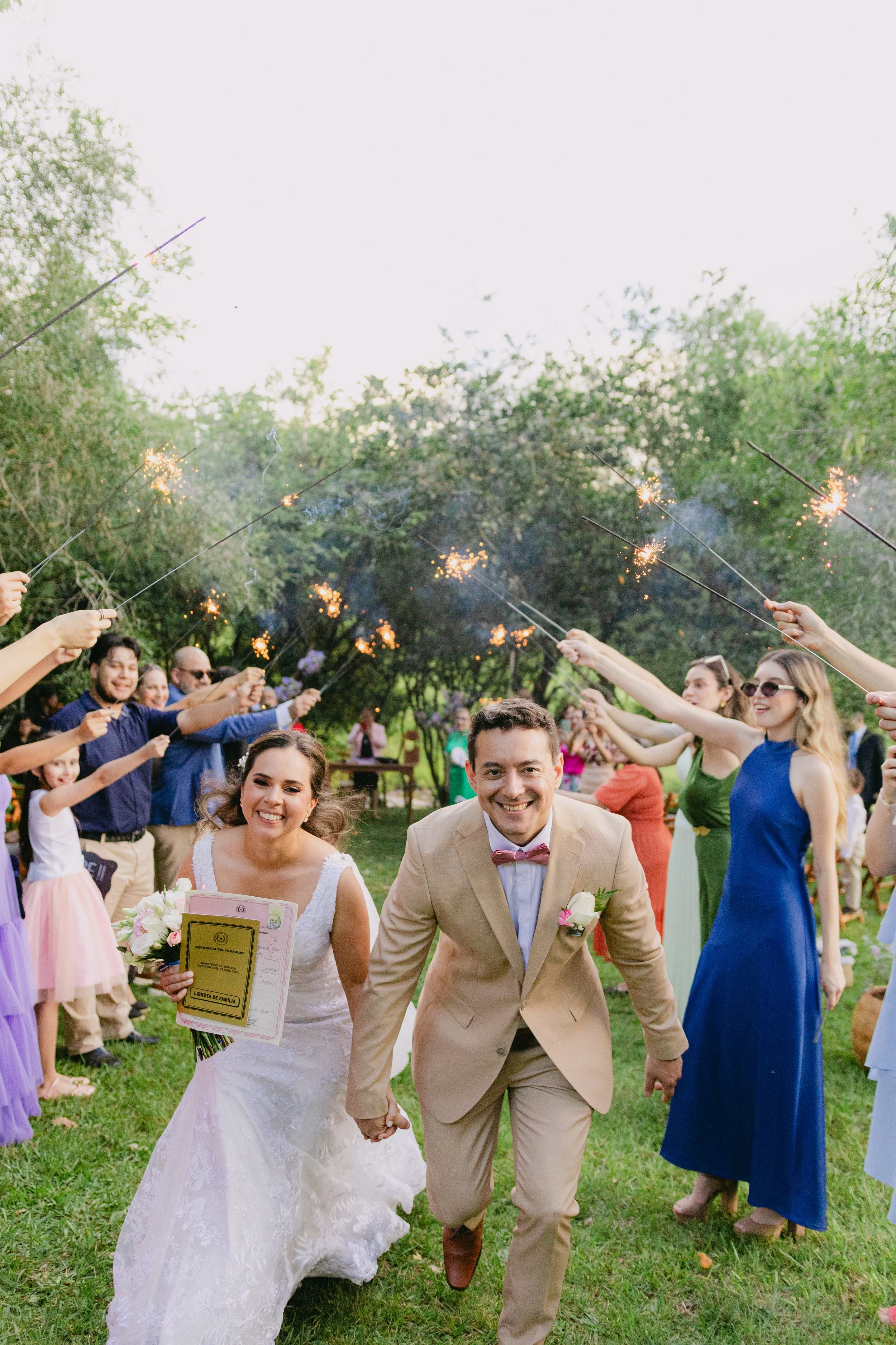 Pareja de novios en su boda, rodeados de amigos y familiares que les felicitan con bengalas en un jardín