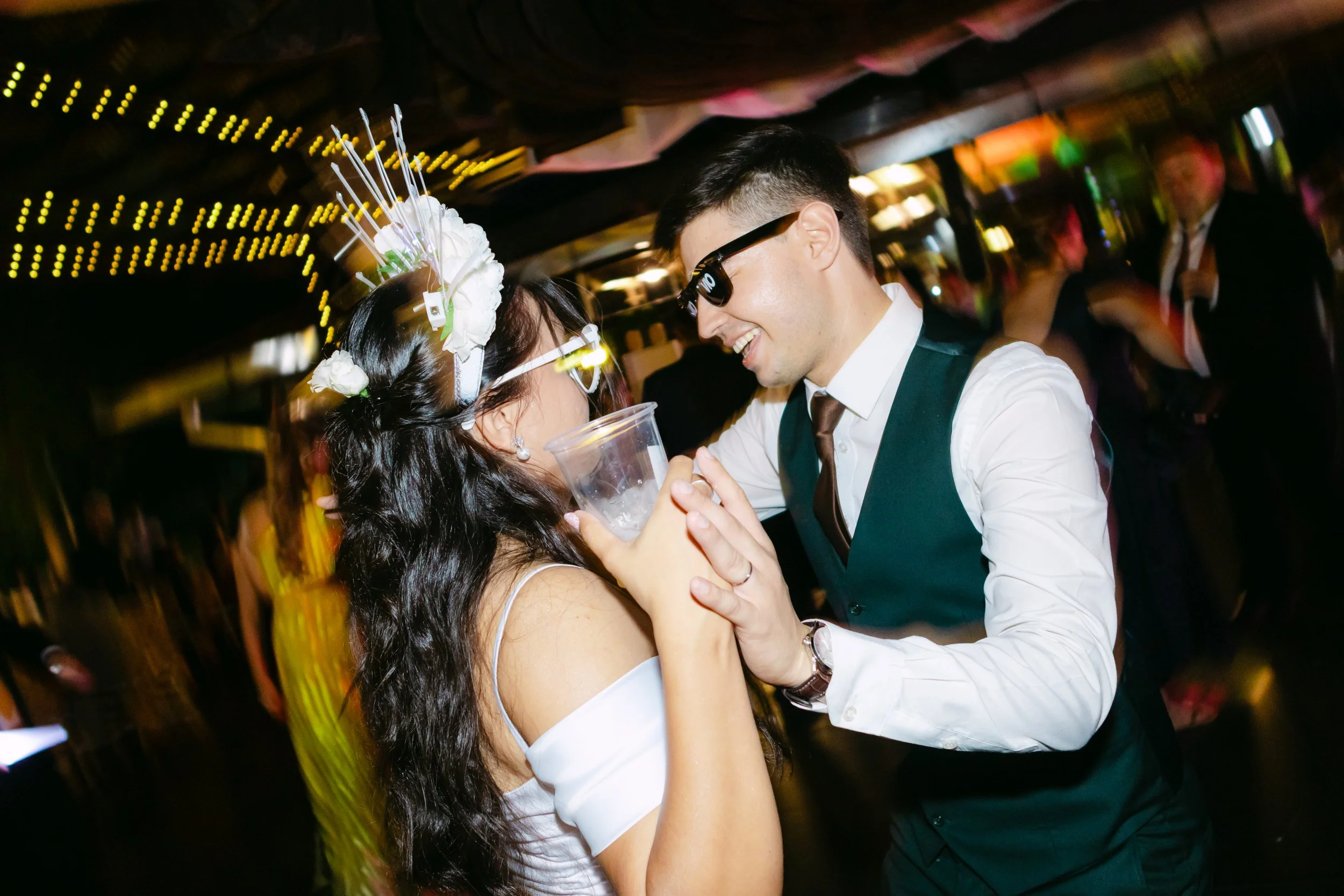 Una pareja bailando y divirtiéndose en una fiesta, rodeada de otros invitados en el fondo, en un lugar con iluminación festiva y colorida.