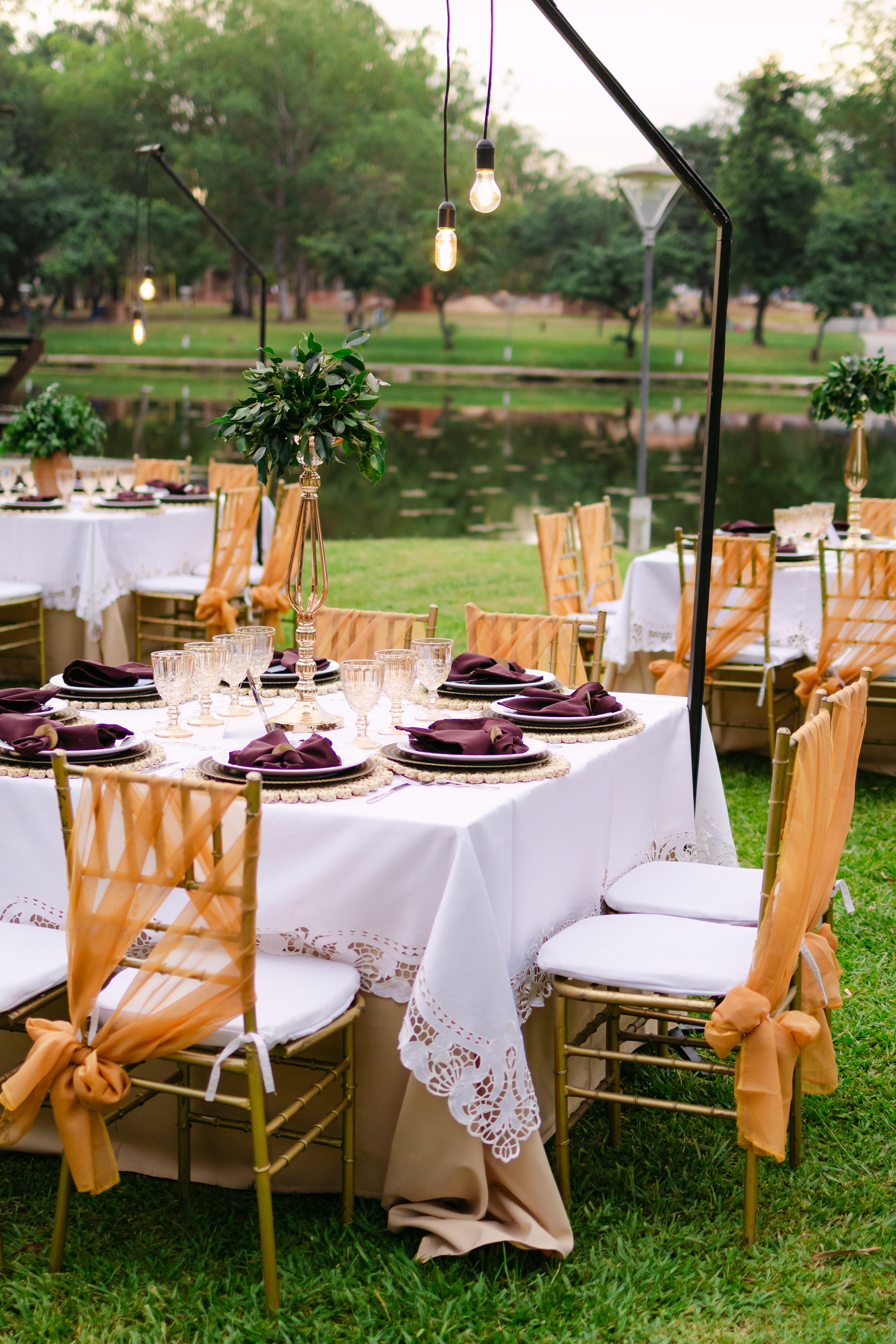 Una mesa decorada para evento al aire libre, con mantelería blanca, vasos y platos de cristalería, sillas doradas con decoraciones de tela anaranjada, centros de mesa con plantas verdes, y luces colgantes, con un fondo de árboles y un lago.
