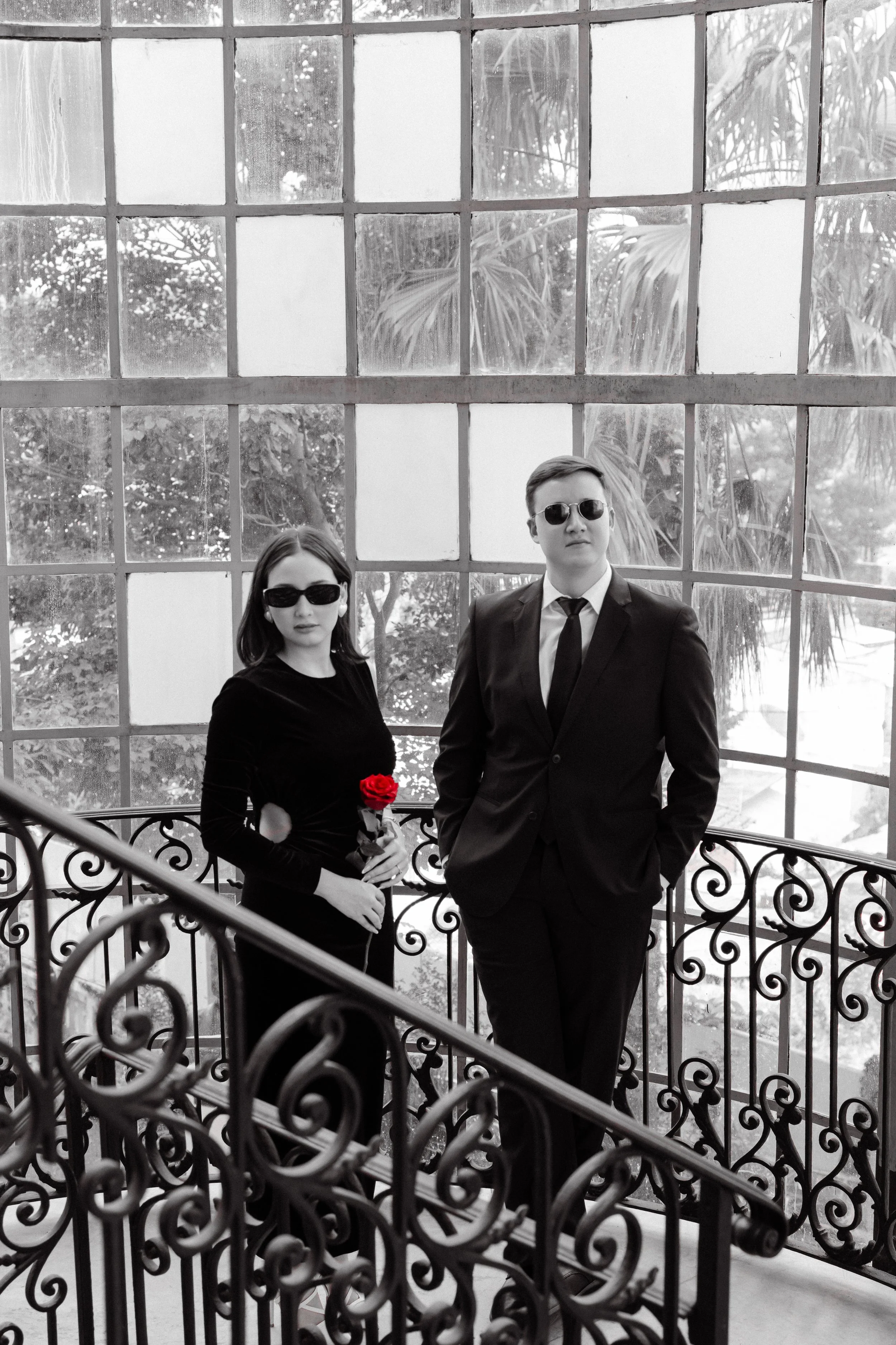 Hombre y mujer vestidos con trajes formales en un lugar con vitrales grandes, la mujer sostiene una rosa roja.