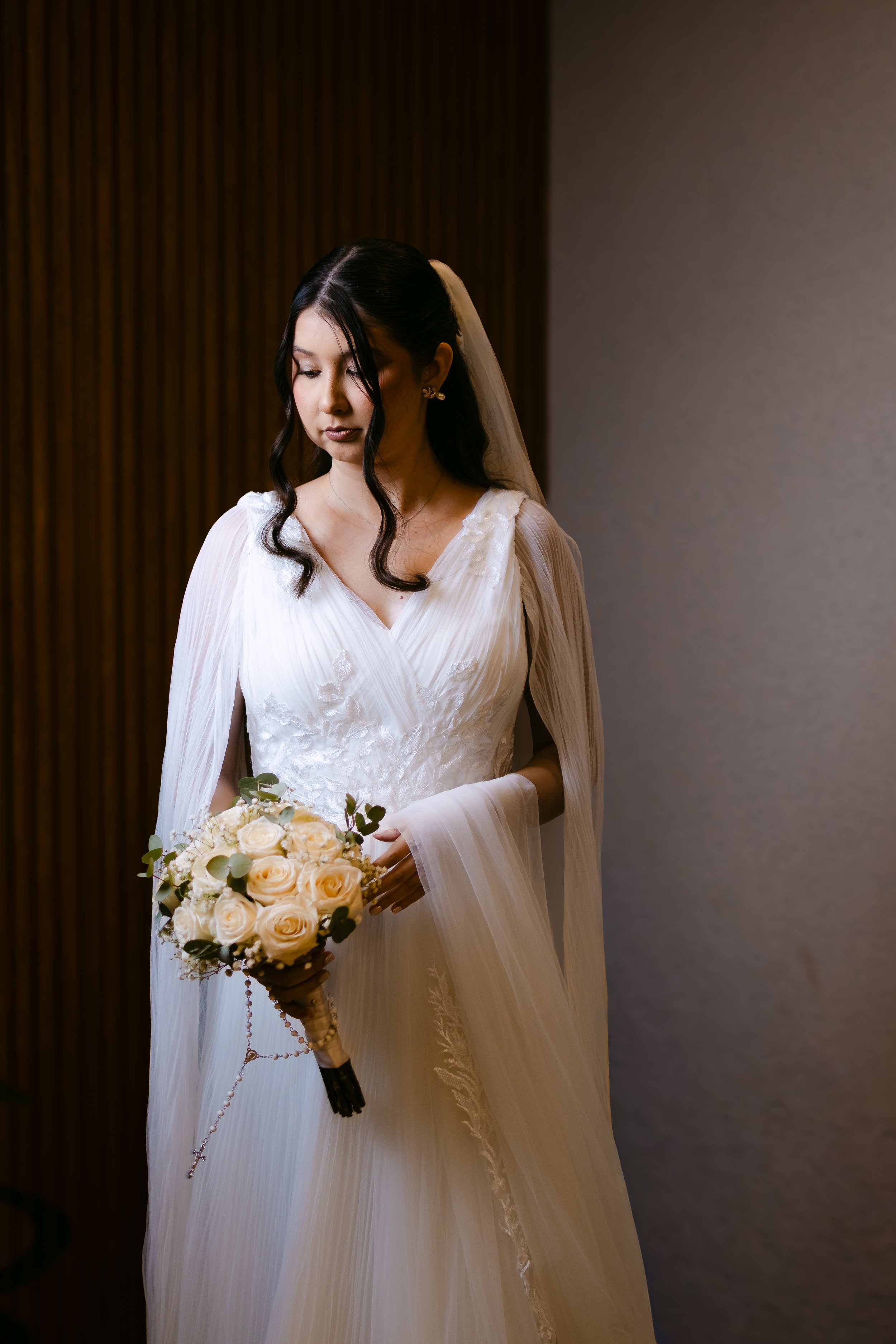 Mujer vestida de novia en un salón, sosteniendo un ramo de rosas blancas.