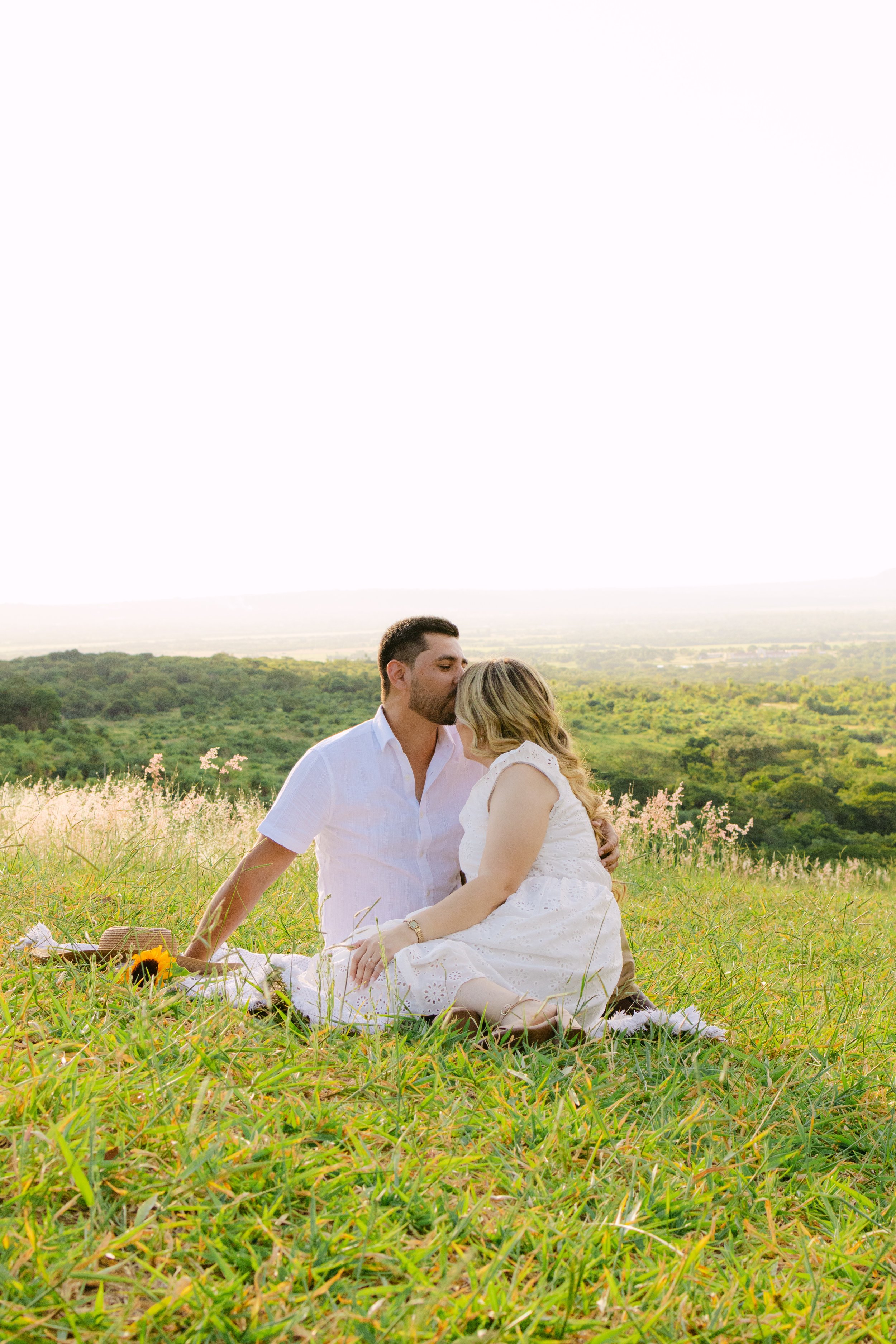 Una pareja sentada en un campo de hierba con flora silvestre, compartiendo un beso bajo la luz del atardecer con un fondo de llanura y vegetación.