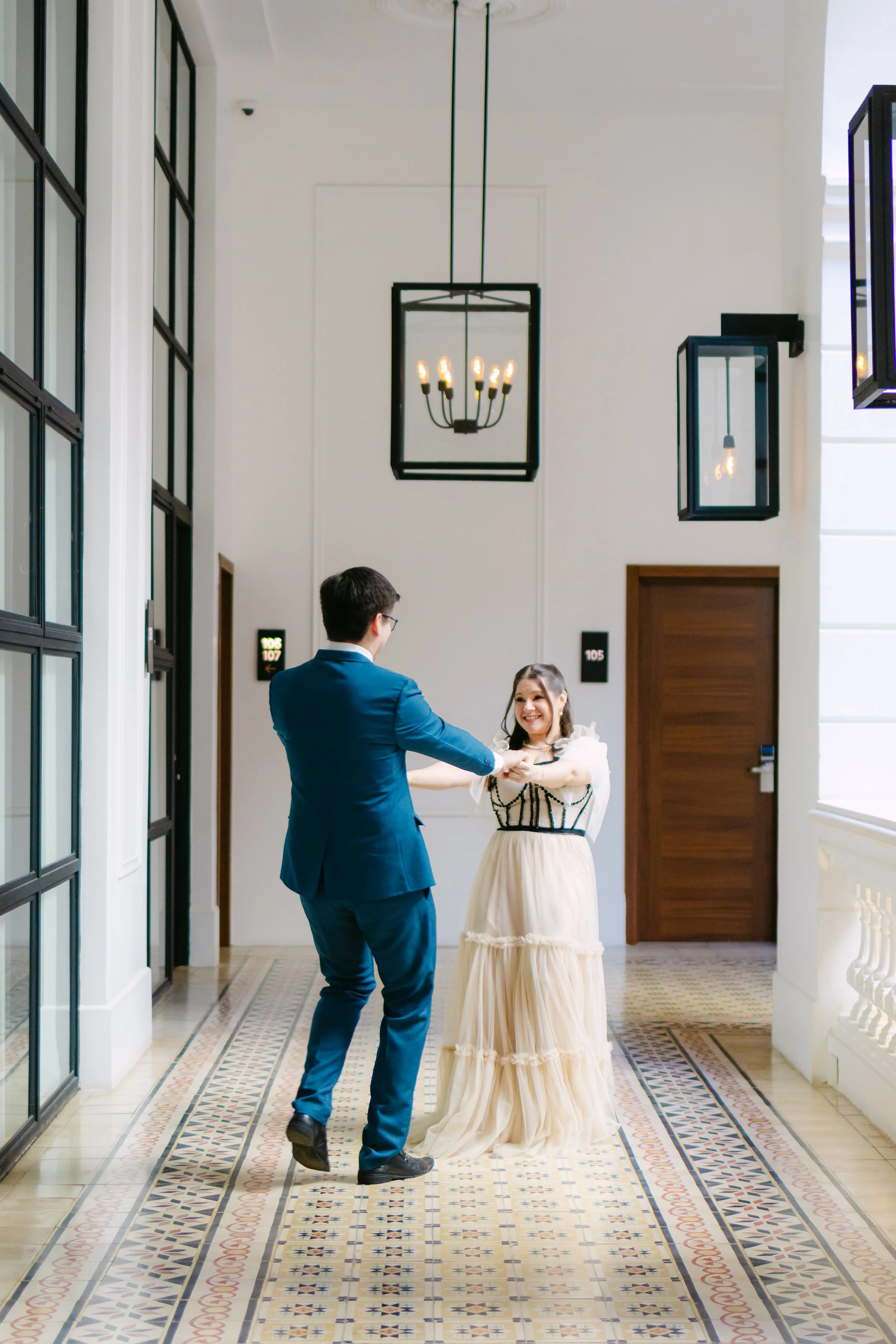 Una pareja bailando en un pasillo elegante con baldosas decorativas y lámparas de techo, con puertas y ventanas grandes.