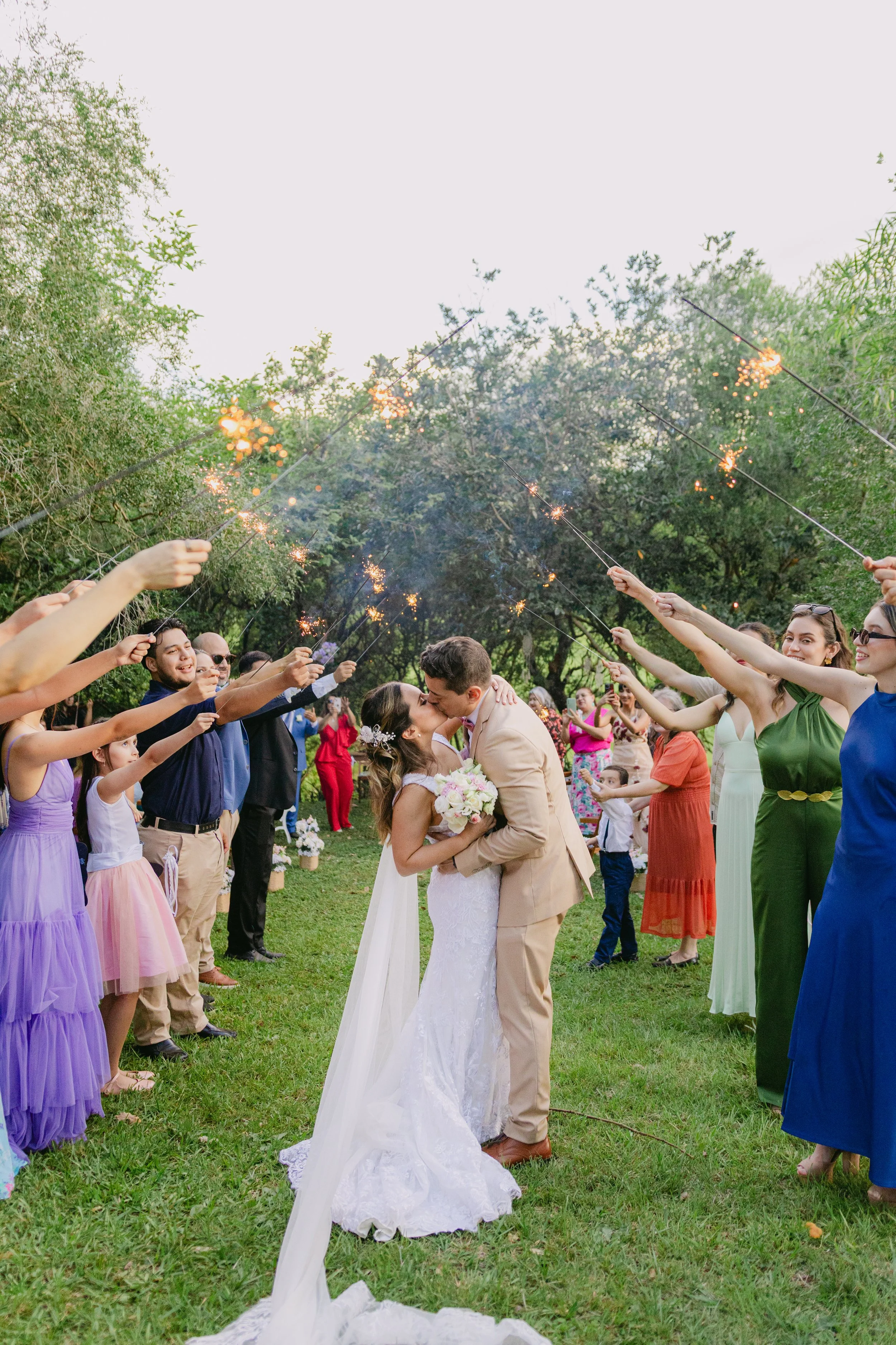 Una pareja de novios se besa en una ceremonia al aire libre con invitados lanzando bengalas en un entorno natural.