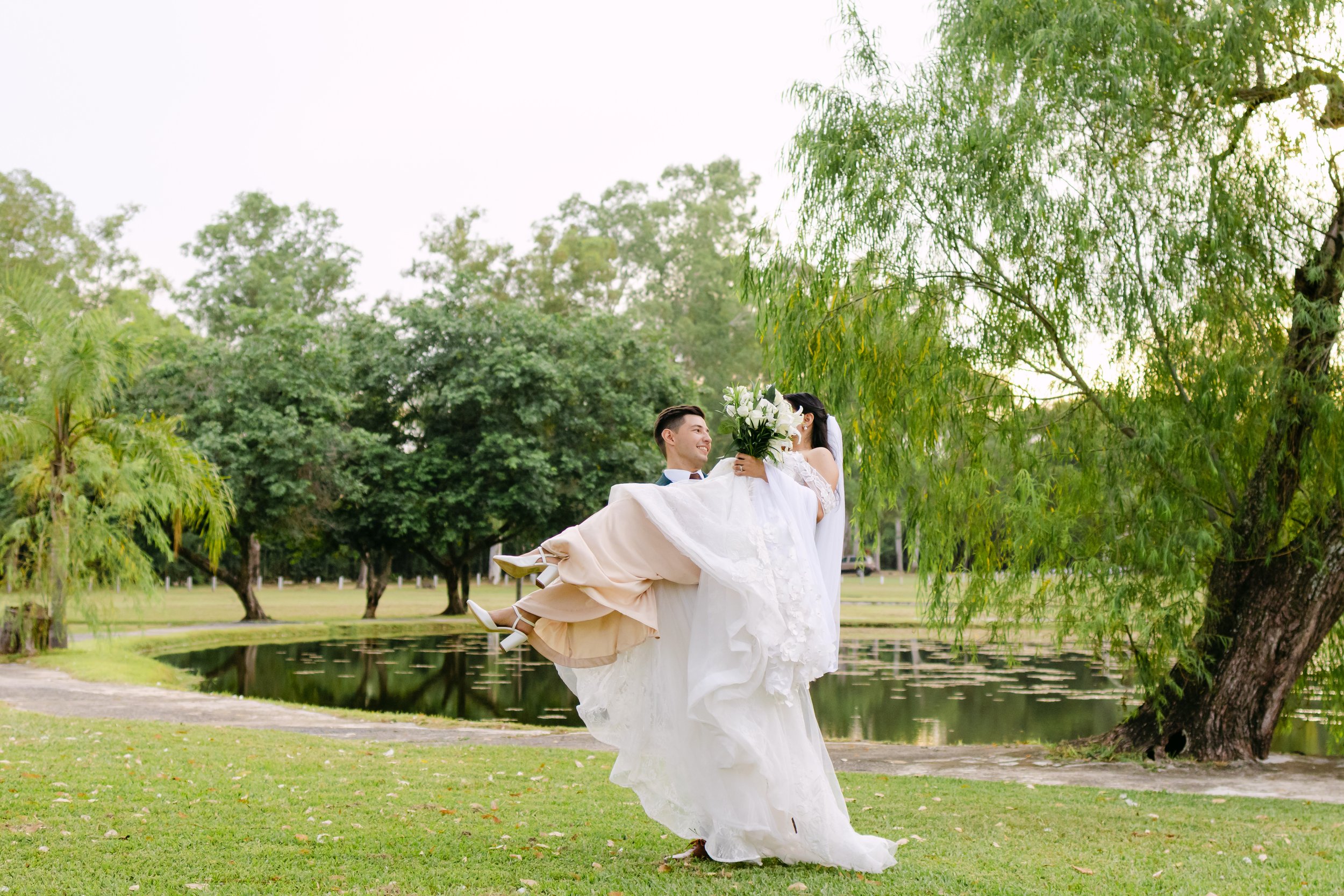 Una pareja de recién casados en un parque, la novia sostiene un ramo de flores blancas y el novio la levanta en brazos, rodeados de árboles y un lago.