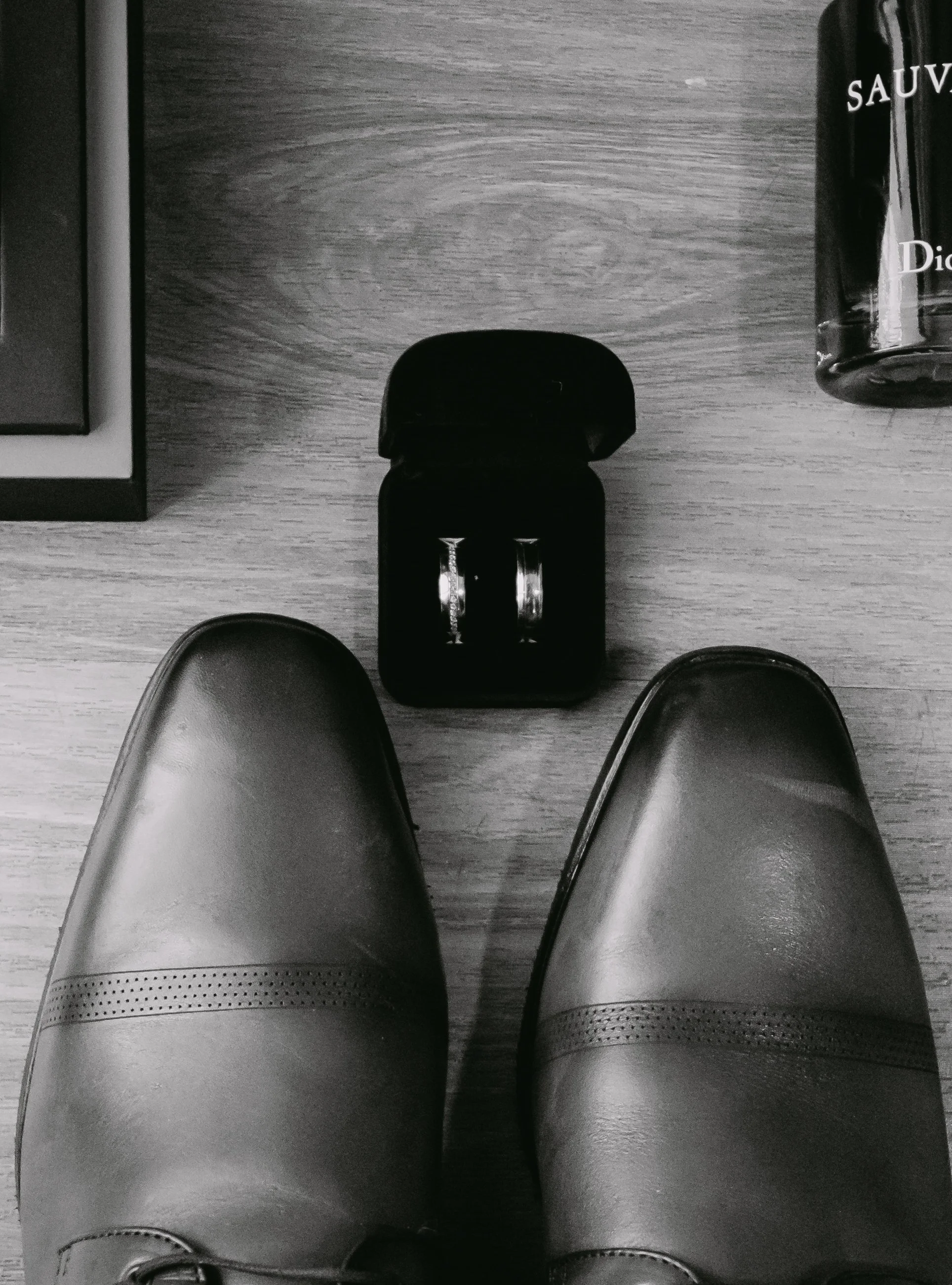 Vista desde arriba de unos zapatos negros de vestir, un estuche con anillos en el centro, una botella de perfume o colonia y una caja en la esquina superior izquierda sobre una superficie de madera.