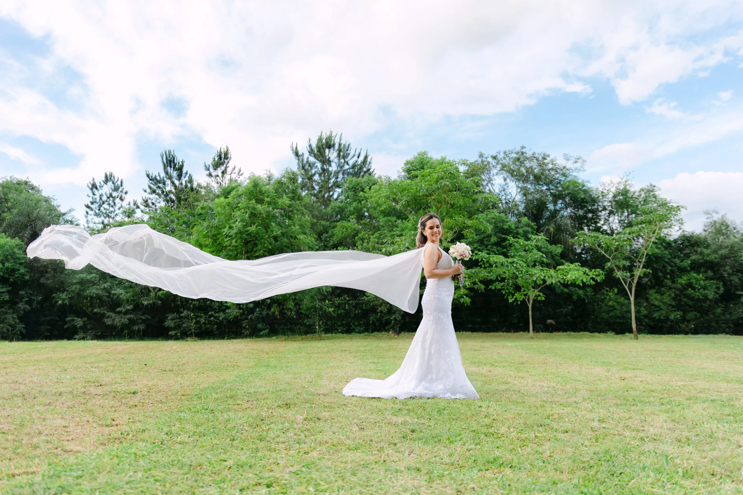 Una novia con vestido de boda largo en un campo con árboles verdes y cielo azul con nubes, sosteniendo un ramo de flores y con la cola del vestido extendida detrás de ella.