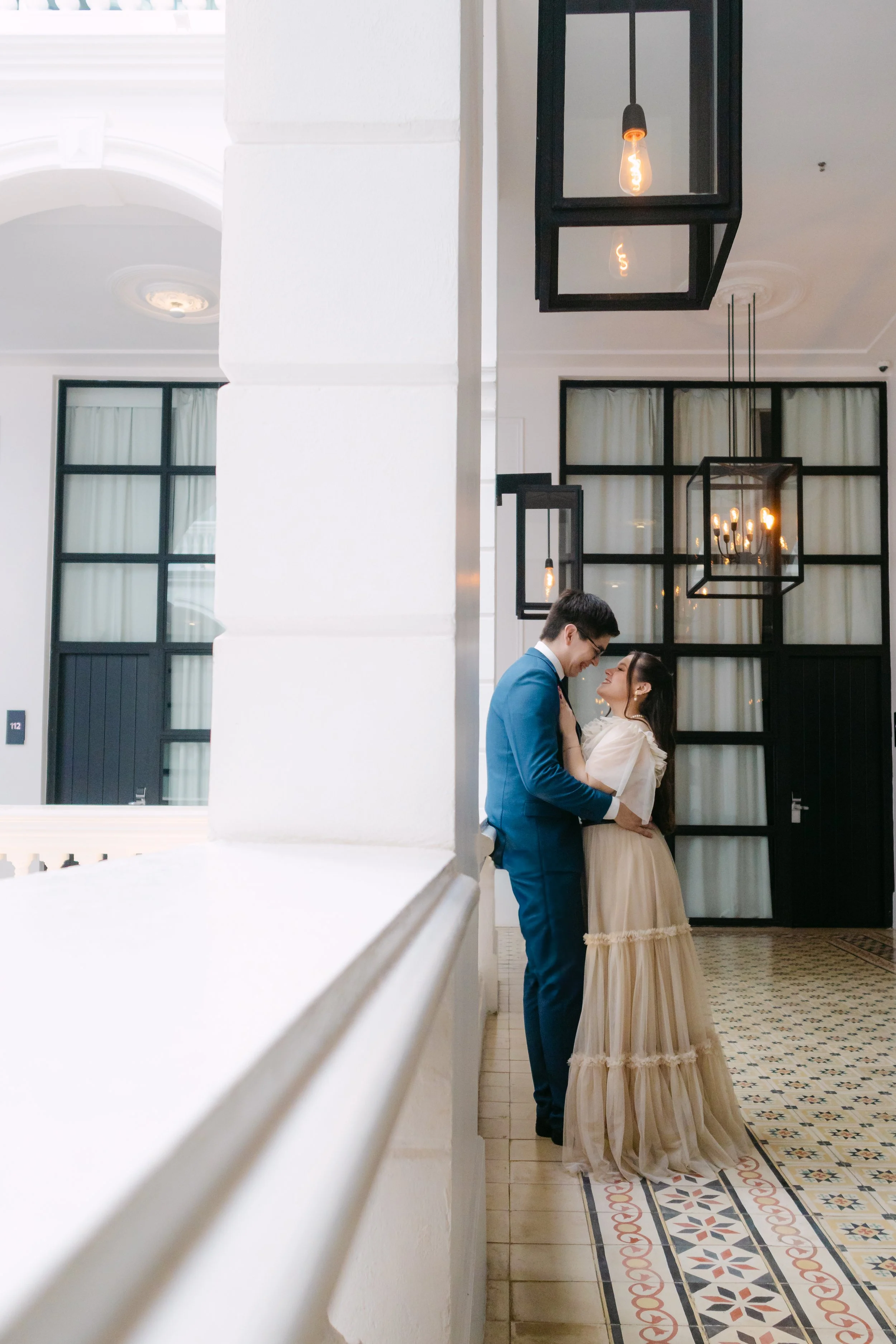 Una pareja vestida formalmente, un hombre en traje azul y una mujer en vestido largo beige, compartiendo un momento romántico dentro de un espacio elegante con iluminación cálida y paredes blancas.