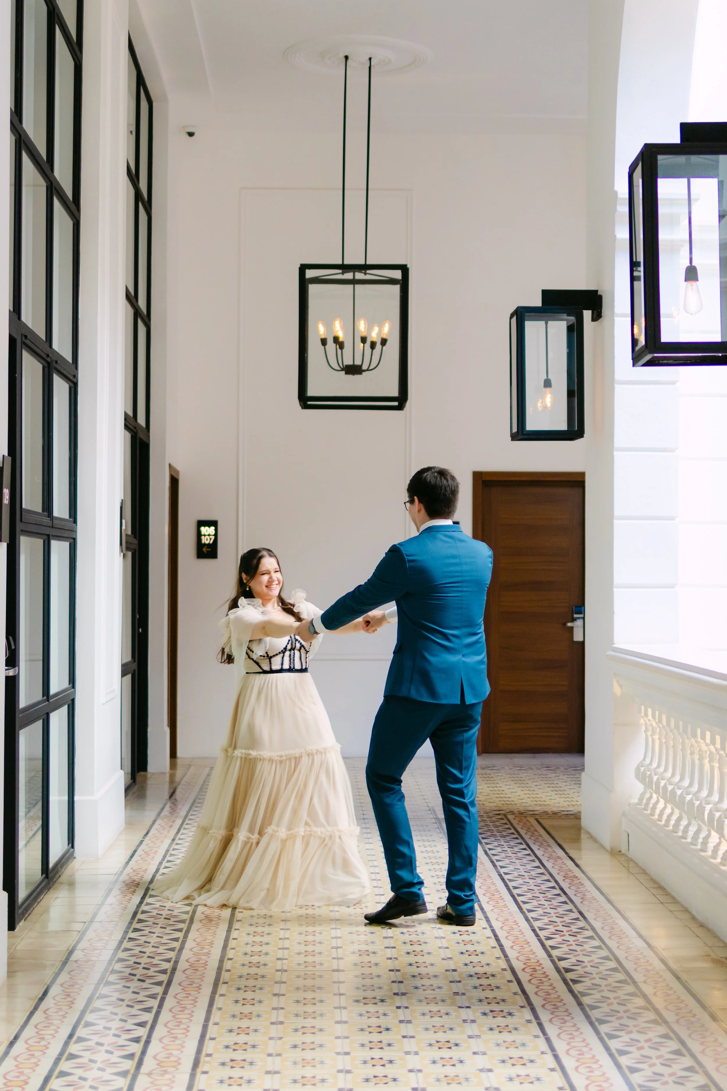 Una pareja bailando en un pasillo elegante con suelo de mosaico y grandes ventanas con marcos negros, decorado con arañas en el techo y paredes blancas.