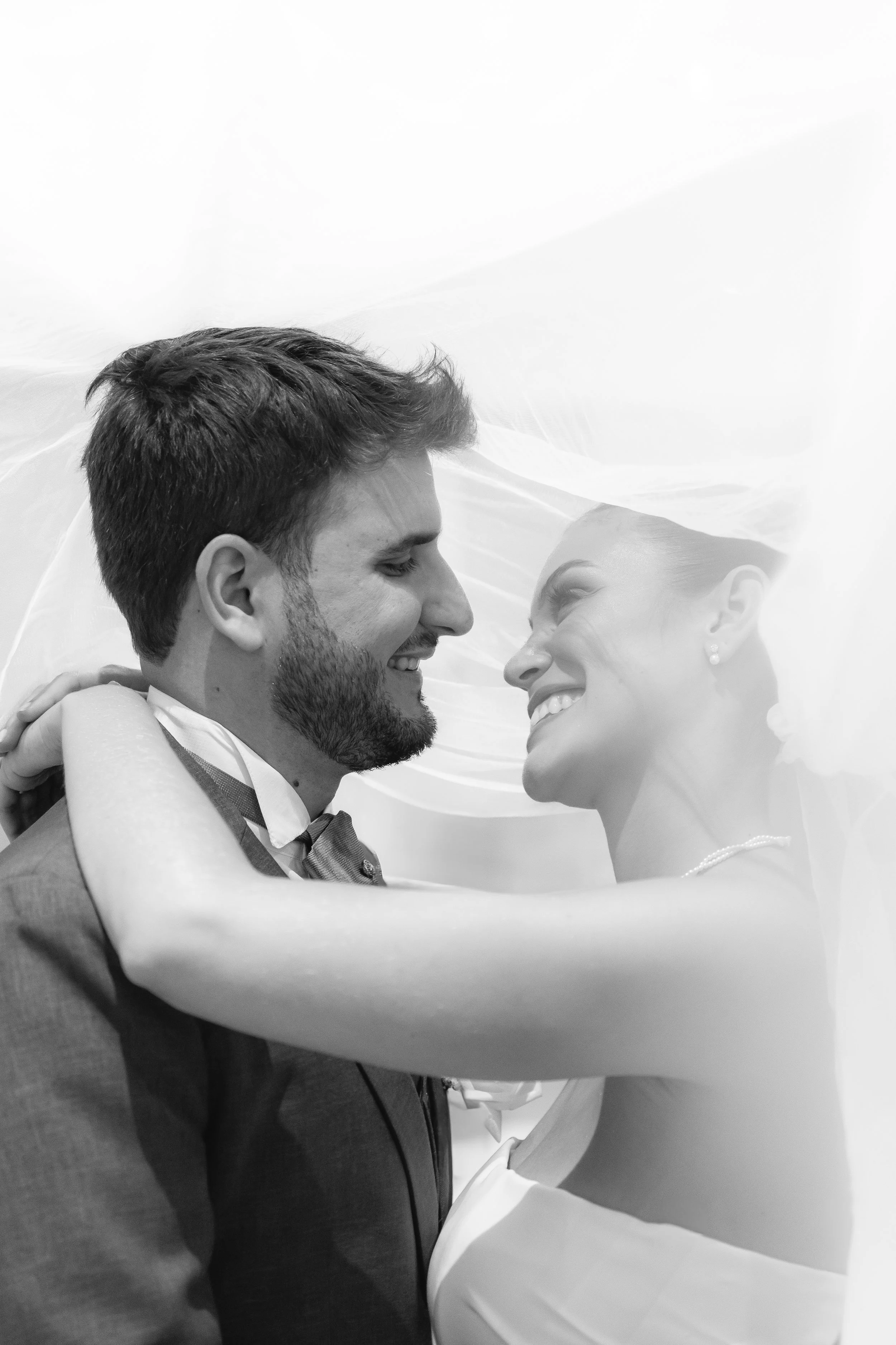 Una pareja de novios sonriente en su boda, abrazándose y mirándose con cariño, en un escenario con velo de novia blanco en blanco y negro.