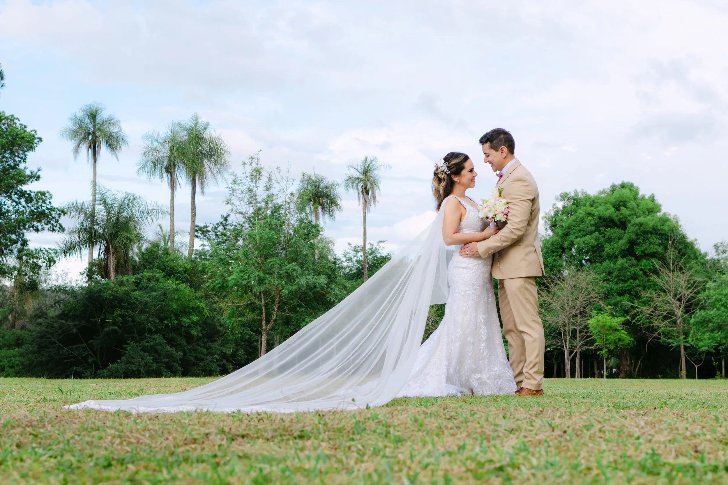 Pareja de recién casados en un campo verde, con árboles y palmas al fondo, ella con vestido de novia y él con traje beige, ambos mirando sonrientes y abrazados, ella con ramo de flores.