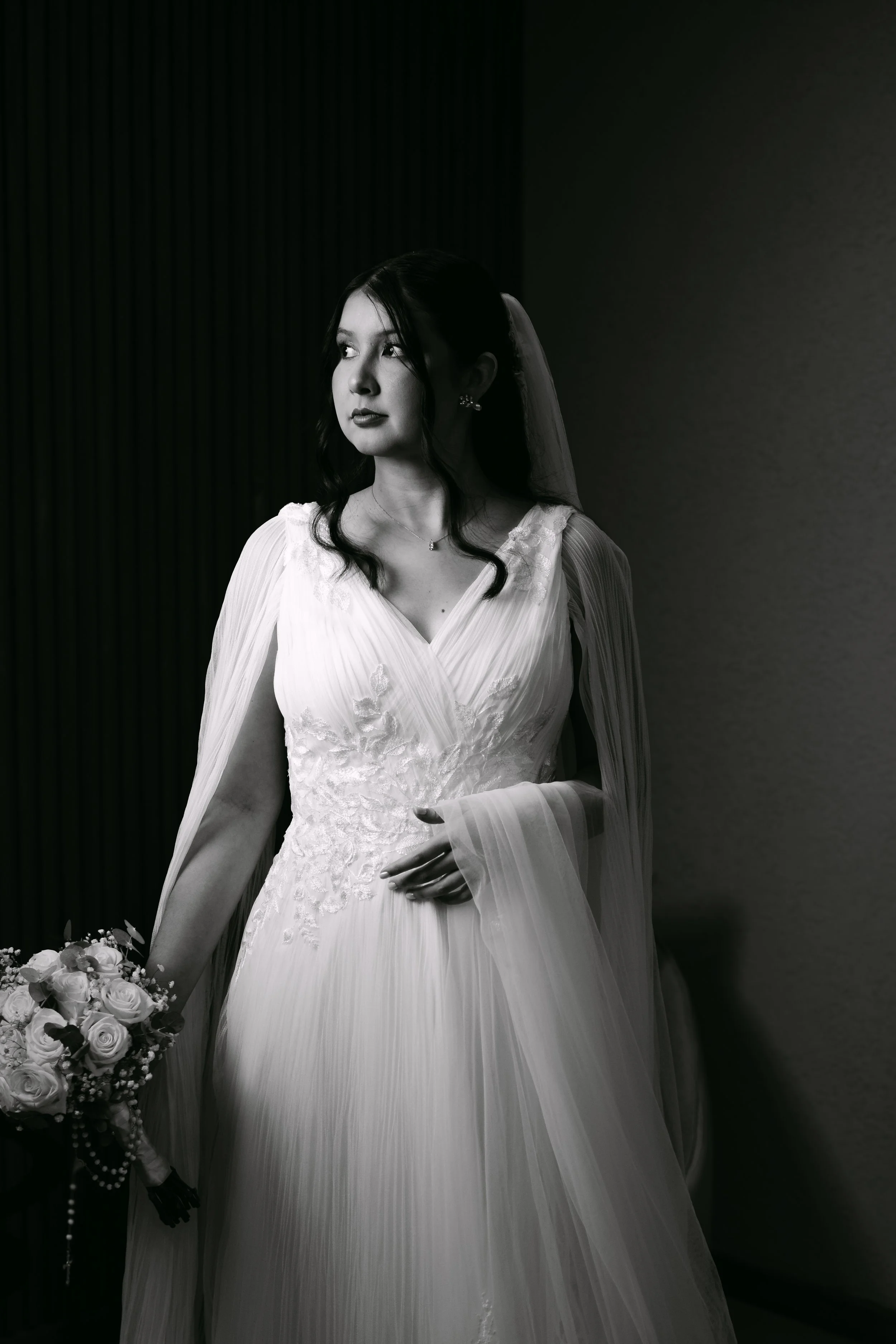 Mujer en vestido de novia en blanco y negro, sosteniendo un ramo de flores, de pie cerca de una pared, mirando hacia la izquierda.